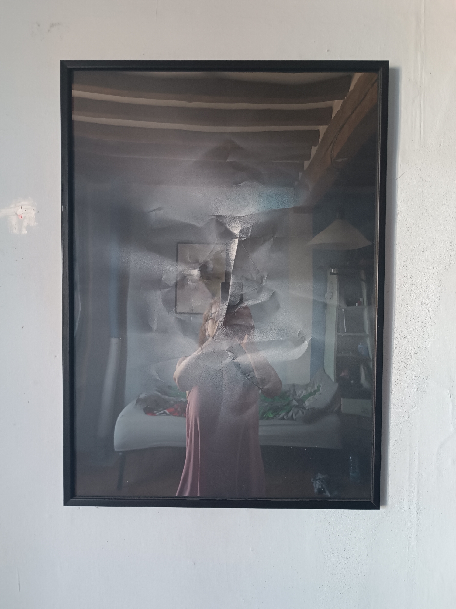 The image shows a framed photograph hanging on a light grey wall. The photograph depicts a partial reflection of a person wearing a reddish dress, standing in a room. The room contains a bed or sofa, shelves, and a hanging lamp. The image is partially obscured by what appears to be crumpled and torn paper, creating an abstract effect. The presence of exposed beams in the upper part of the reflection suggests the setting is an interior with architectural features common in older structures. Without additional information, the specific location in Viadana, Italy, cannot be determined.