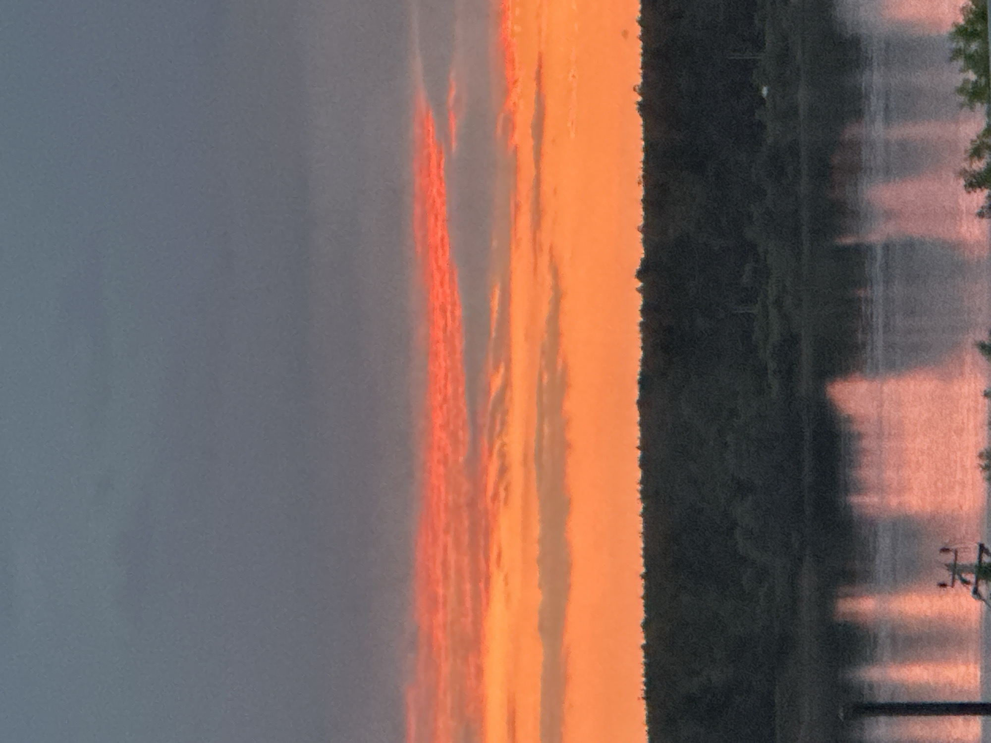 The image captures a serene sunset over the Kama River in Chaikovsky, Russia. The sky is a vibrant canvas of color, with streaks of bright orange and red hues illuminating the horizon. The river itself reflects the warm glow of the sunset, creating a shimmering, almost ethereal effect on its surface. A dense forest lines the riverbank, its dark silhouette contrasting with the fiery sky. The scene is tranquil and visually striking, evoking a sense of peace and natural beauty.