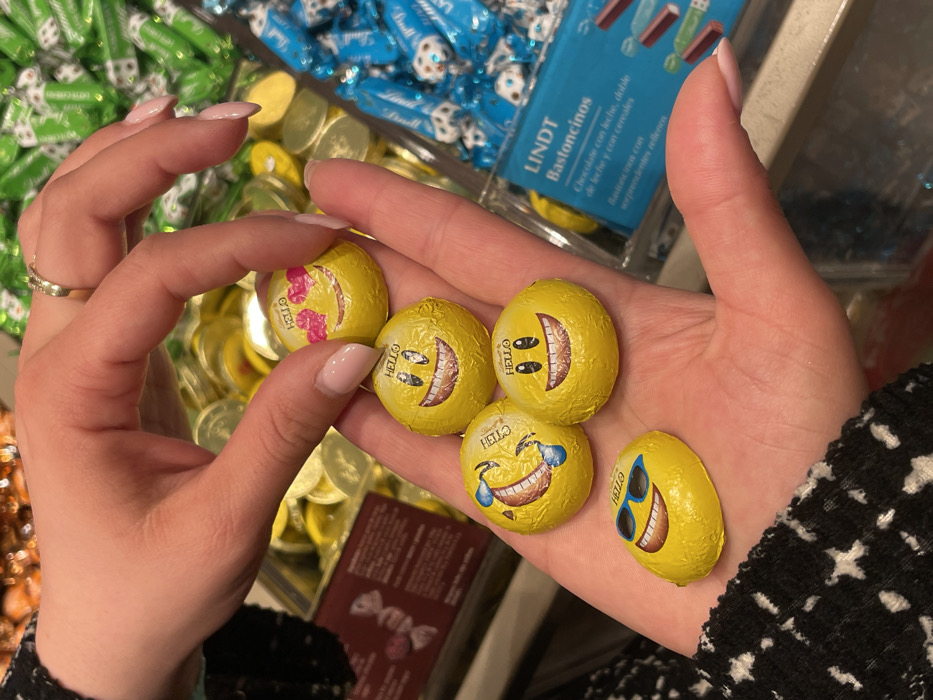 A close-up photograph captures two hands interacting with five emoji-themed chocolate candies within a retail environment in Marratxí, Spain. The upper hand, adorned with a gold ring on the ring finger and light-colored nail polish, delicately holds one circular yellow-foiled chocolate depicting a "heart eyes" emoji and the text "HELLO." The lower hand, open with its palm up and also displaying light-colored nail polish, cradles four additional yellow-foiled chocolates. These include one circular "grinning face with smiling eyes" emoji, one circular "face with tears of joy" emoji, another circular "grinning face with smiling eyes" emoji, and an oval-shaped "smiling face with sunglasses" emoji, all bearing the "HELLO" text. In the blurred background, various bins or displays of wrapped candies are visible, including green-wrapped items to the upper right, gold-foiled circular chocolates, and a transparent container on the right holding blue-wrapped Lindt Bastoncitos. A dark red sign with white text is partially visible on the left side of the frame.