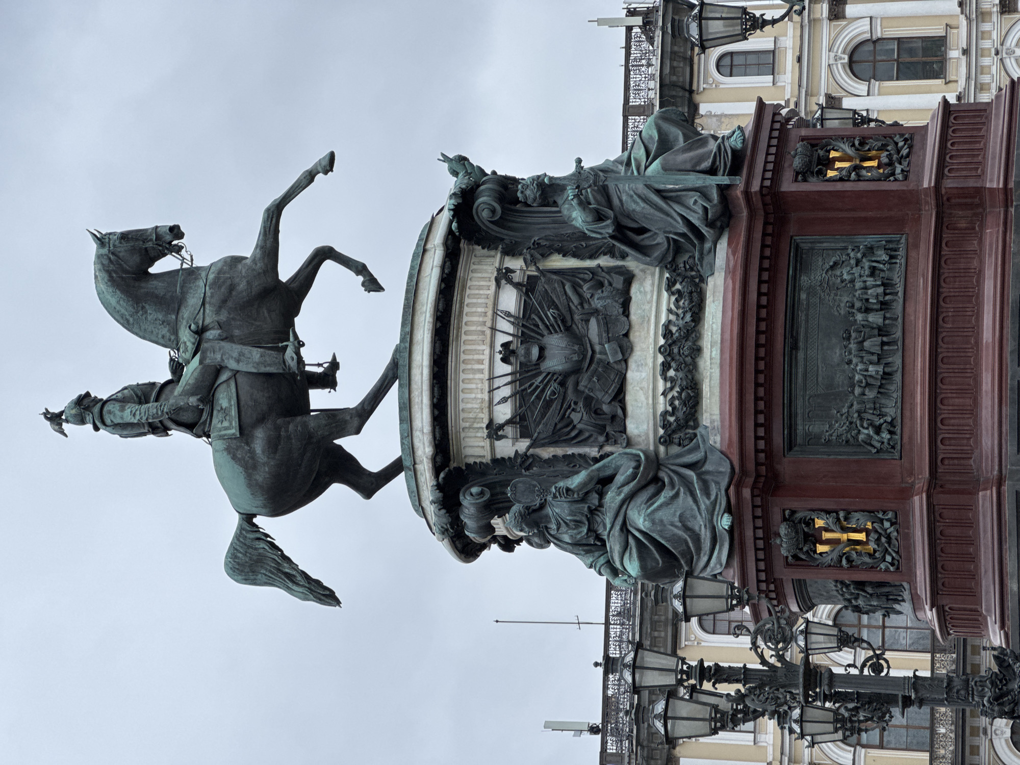 This media file presents a grand equestrian statue under an overcast sky. The central subject is a large bronze statue depicting a male figure, likely a monarch or military leader, mounted on a dynamically rearing horse. The horse is balanced on its hind legs, with its front legs raised high. The rider is dressed in military attire, including a helmet, and extends his right arm forward, holding a small, indistinct object. The bronze has acquired a greenish patina over time.
The statue rests on an elaborate, multi-tiered pedestal. The lower section of the pedestal is made of a polished, dark red-brown stone, while the upper sections feature detailed bronze work and lighter-colored stone. Allegorical figures are seated at the corners of the pedestal, and intricate bas-reliefs depict historical or symbolic scenes, including one showing a gathering of figures and another with military trophies. Notable gilded monograms, possibly "N I" (for Nicholas I), crowned and set within ornate frames, are visible on the red stone section of the pedestal.
In the background, parts of a large, classical-style building are visible, characterized by light-colored walls, arched windows, and decorative architectural details. Ornate black metal streetlights with multiple lanterns are also prominent in the lower part of the image. The setting, with its monumental sculpture and grand architecture, strongly suggests a significant historical location, consistent with Saint Petersburg, Russia. The overcast weather contributes to a soft, diffuse light across the scene.