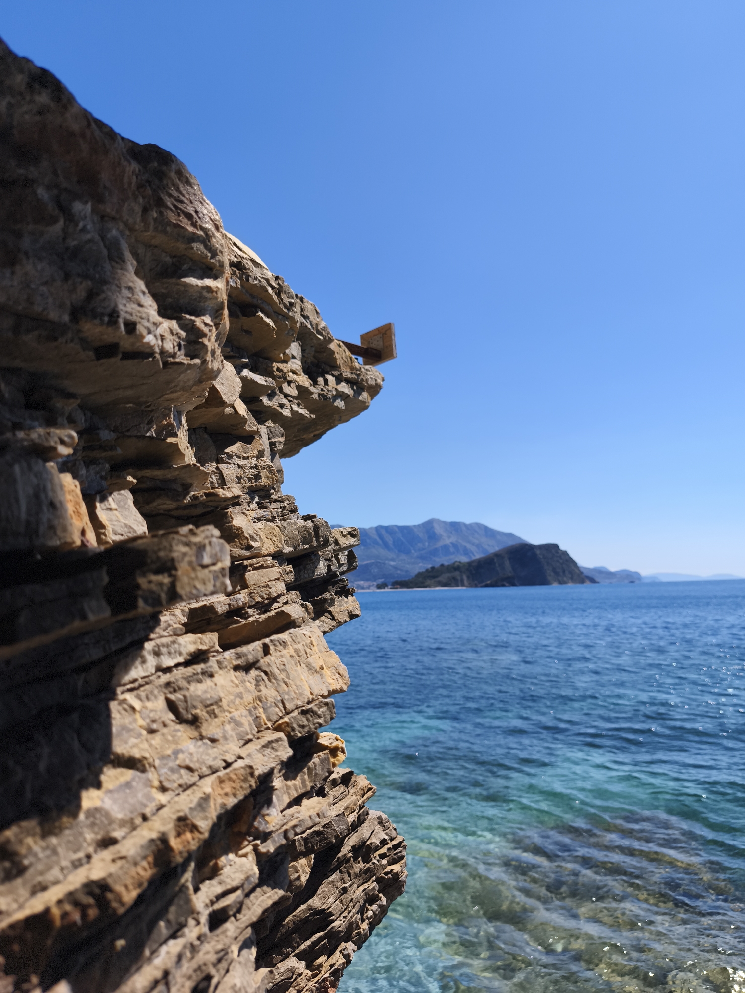 The image depicts a rocky cliff face overlooking a body of water. The cliff is composed of layered sedimentary rock with variations in color and texture. A rectangular structure, potentially artificial, is situated on the cliff's edge.
In the background, a landmass with mountainous terrain is visible across the water. The water itself displays varying shades of blue and turquoise. The sky is clear and blue.
The location appears to be coastal, specifically in Budva, Montenegro.