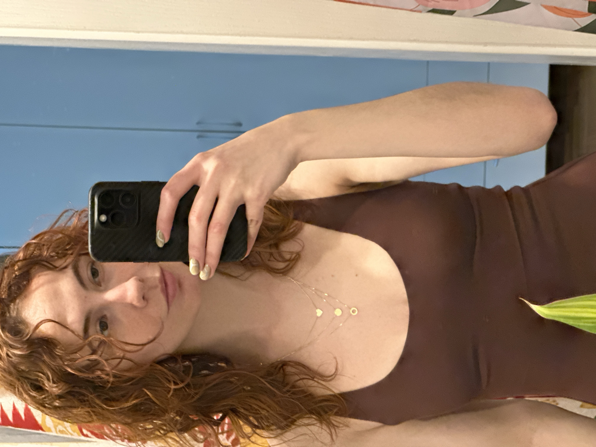 A woman with reddish-brown curly hair is taking a selfie in a mirror. She is wearing a dark brown sleeveless top and a delicate gold necklace with small charms. Her nails are painted with a light green and gold design. She is holding a black smartphone with multiple camera lenses. The reflection shows a blue cabinet or wall behind her, and a plant with green and yellow variegated leaves is partially visible on the right side of the frame. The location is likely indoors, but no specific details suggest Novi Sad, Serbia.