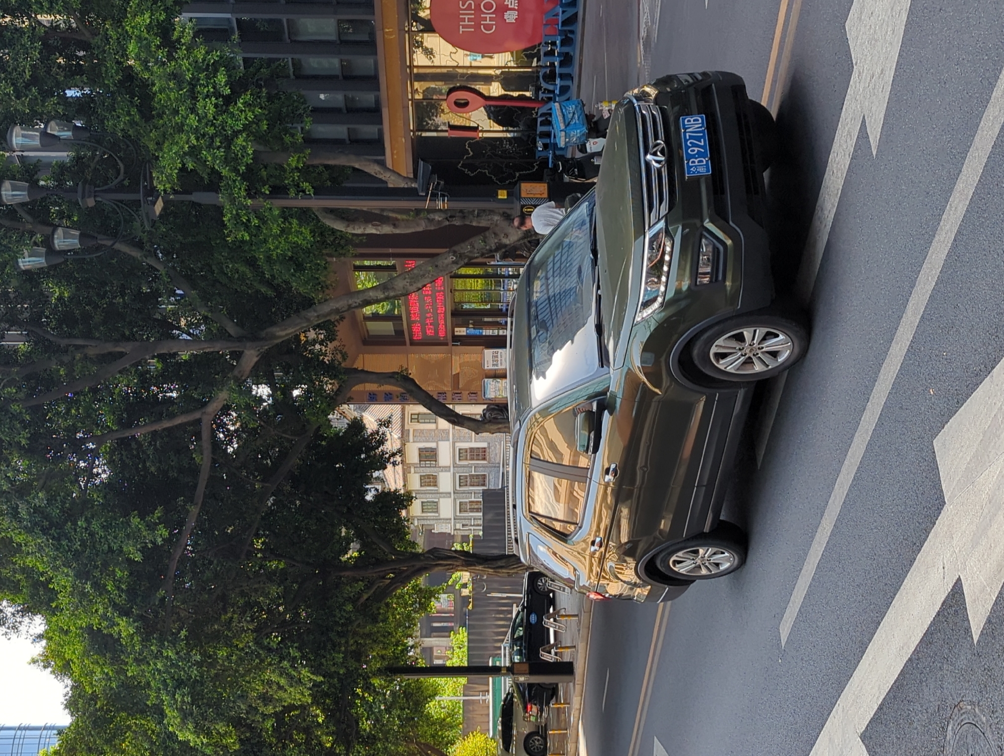 In Yuzhong District, China, a dark green SUV is traveling on an asphalt road marked with white directional arrows. The vehicle has a license plate that reads "B-927NB". The scene is framed by lush green trees and buildings with traditional Chinese architecture, suggesting an urban environment with established foliage. In the background, several other vehicles are parked along the roadside, and a streetlamp with multiple lights is visible. A person in a white shirt is walking near the sidewalk. The overall atmosphere is that of a bright, sunny day in a bustling city.
