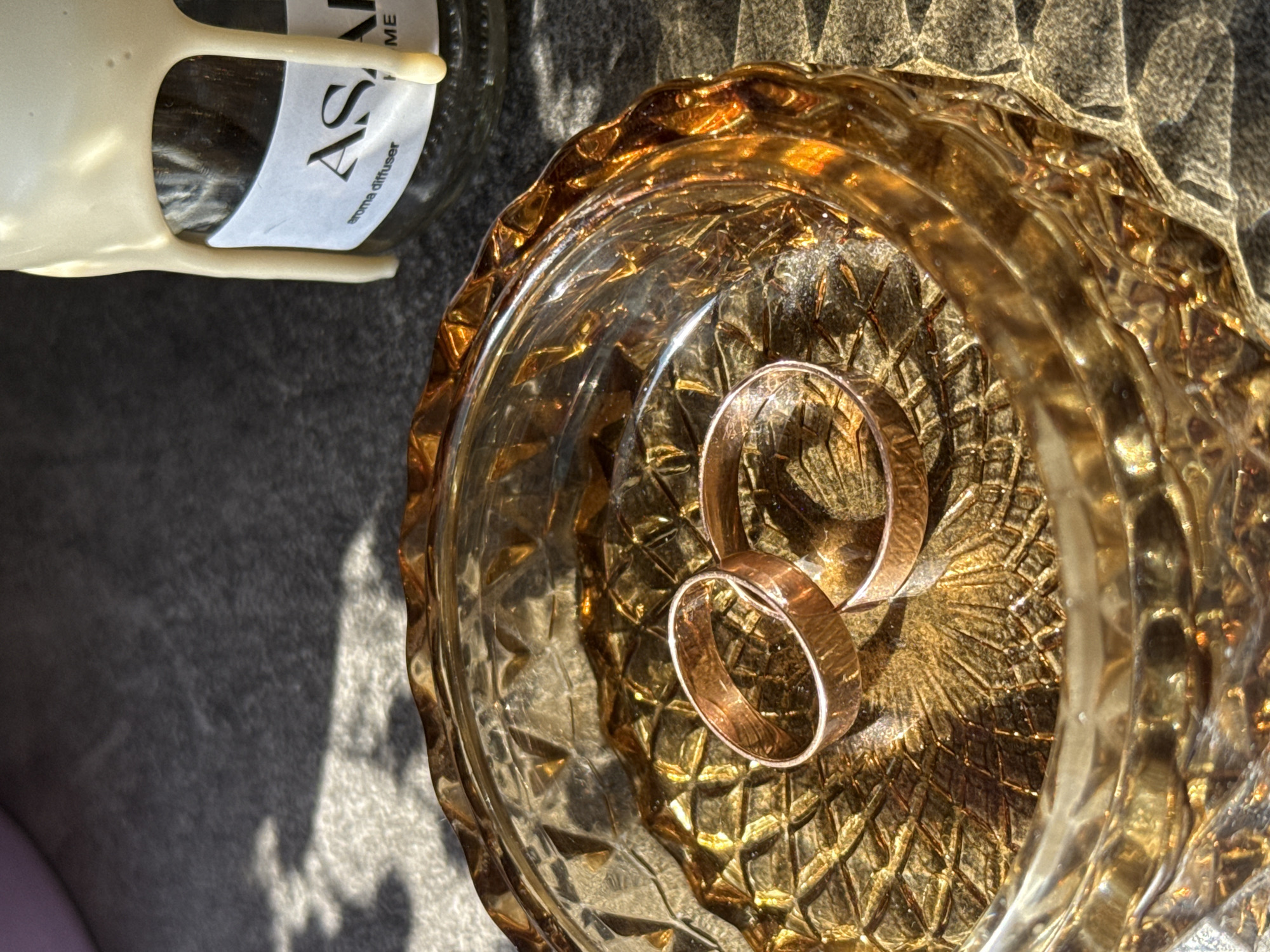 The image is a close-up, still-life photograph of two rose-gold wedding bands nestled in a vintage, amber-colored glass bowl. The bowl features a heavily textured, possibly cut-glass design. A small portion of a white label with the text "Aroma diffuser" is visible in the upper-left corner, adhered to a glass bottle with a white plastic dispensing mechanism. The items are resting on a textured, grey surface. The lighting suggests it is outdoors with bright sunlight. There is no indication of the location being in Petropavlovsk-Kamchatsky, Russia. No people are visible in the image. The interaction is simply the placement of the rings within the bowl for the photograph.