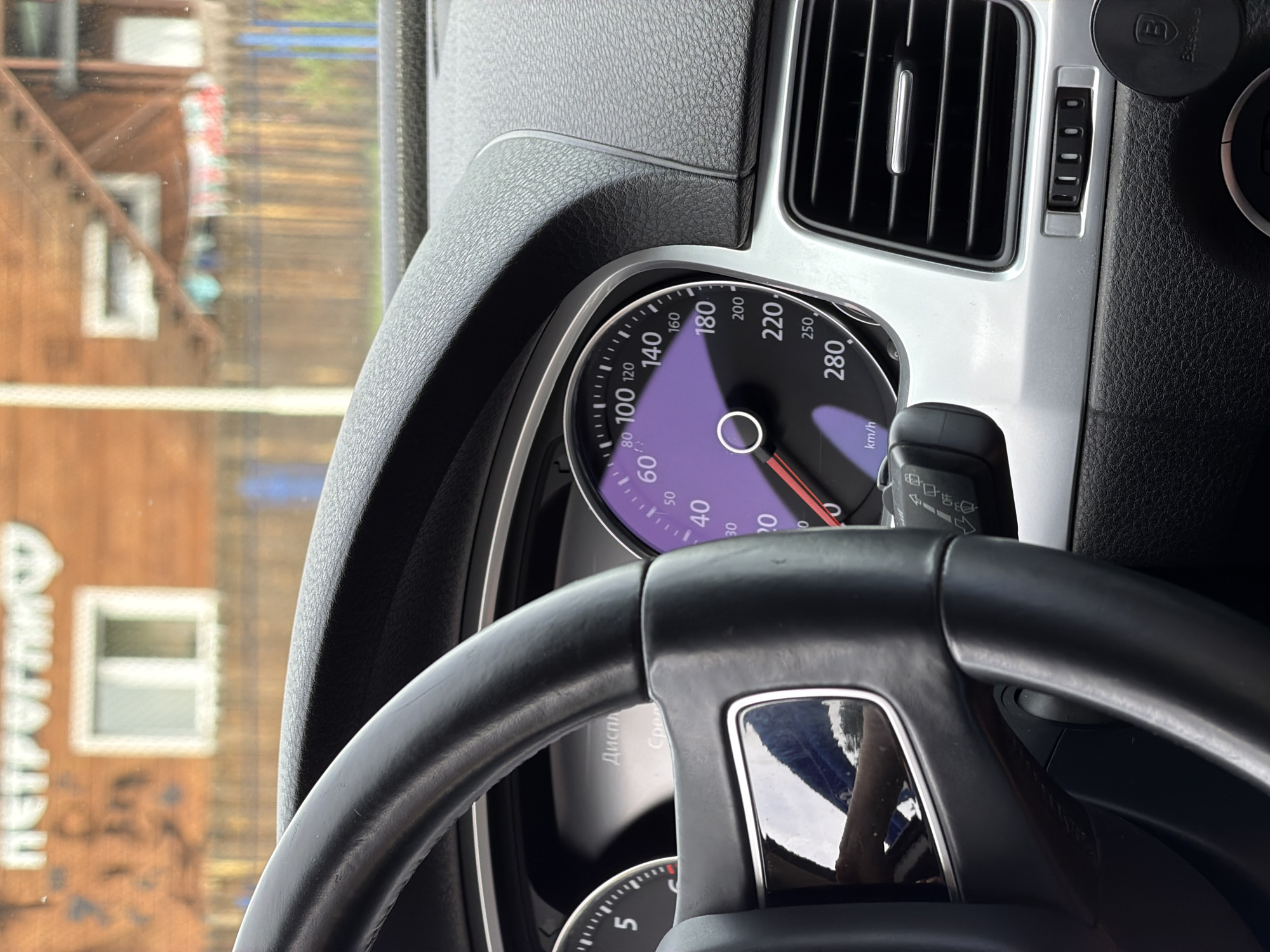 The image, taken inside a vehicle, focuses on the dashboard and steering wheel. A portion of the dashboard, including the speedometer which displays a speed of approximately 40 km/h, is visible. The steering wheel, a black leather-wrapped steering wheel, is prominently featured in the foreground. The background shows a blurred view of a building with wooden siding and a window. Text, "ПЕТРОГЛИФЫ" (petroglyphs), is visible on the building's facade. A sign with the logo of "Baseus" is attached to the lower right of the dashboard, suggesting the presence of a phone mount. The scene is likely set in the city of Катунь, Russia, given the text in Russian.