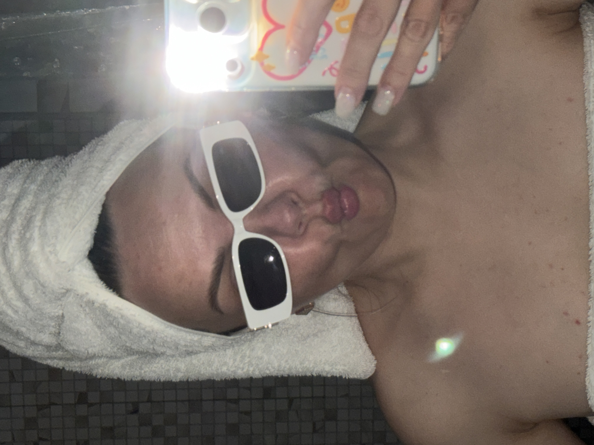 A selfie of a person with a white towel wrapped around their head and wearing white-framed sunglasses with black lenses. The person is pouting and holding a cell phone up to take the photo. Their left shoulder is visible. The location of the photo is not discernible, nor is the city of Votkinsk, Russia.