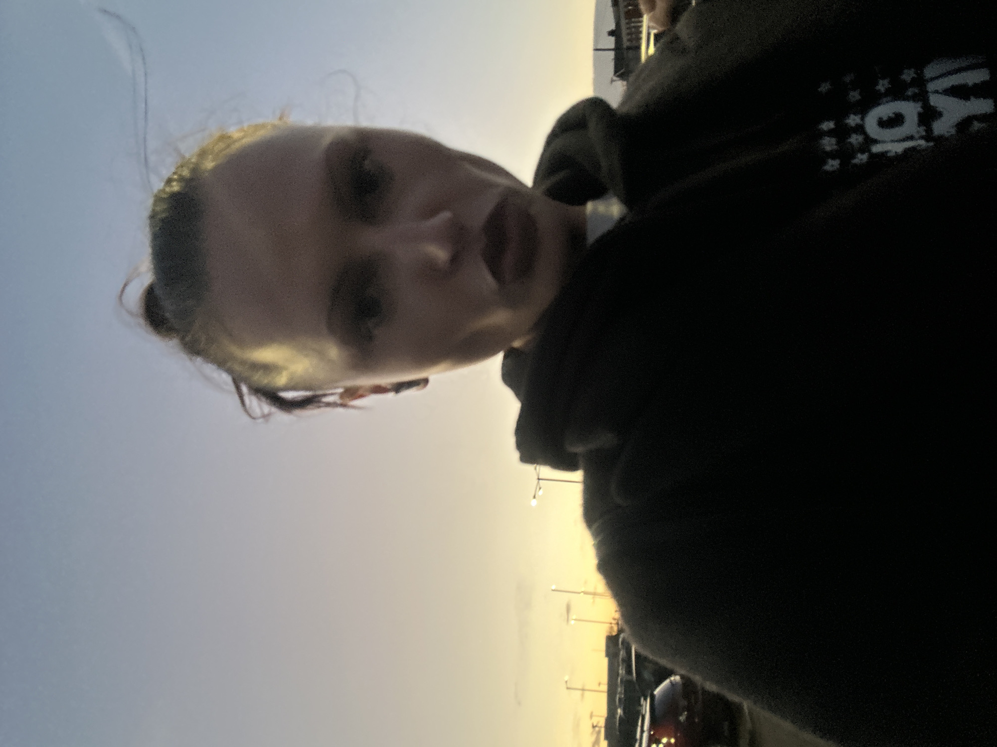 The image displays a female subject, primarily framed from the chest up, positioned centrally and directly facing the camera. She has dark hair pulled back into a bun with loose strands and an earbud visible in her left ear. Her lips are pursed, creating a "duck face" expression. She wears a dark hooded sweatshirt with a light-colored graphic on the right side of the chest, depicting stars and the characters "10:Y". The background reveals an outdoor parking area in Sevenoaks, United Kingdom, during either dawn or dusk, characterized by a sky gradient shifting from orange-yellow at the horizon to light blue overhead. Several tall streetlights with illuminated fixtures are visible, alongside multiple parked vehicles, with only their upper portions or rear lights discernible. No interactions between individuals are observed.