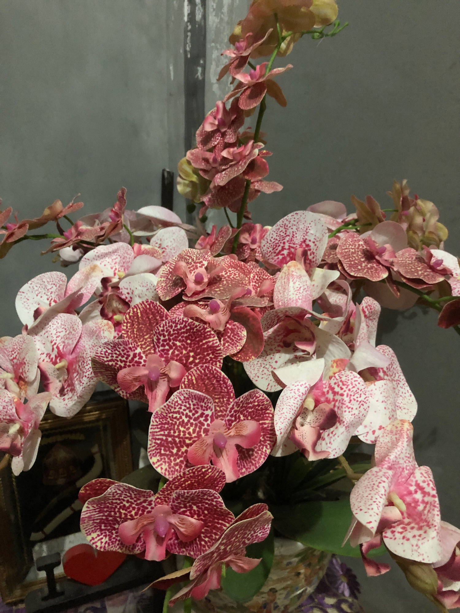 A cluster of artificial pink and white orchids with red speckles is displayed in front of a gray concrete wall. To the left, a framed picture is visible, with a red heart-shaped object and the letter "I" beneath it. The overall impression is of a decorative arrangement. The location of this scene in Lhokseumawe, Indonesia, is not evident from the image itself.