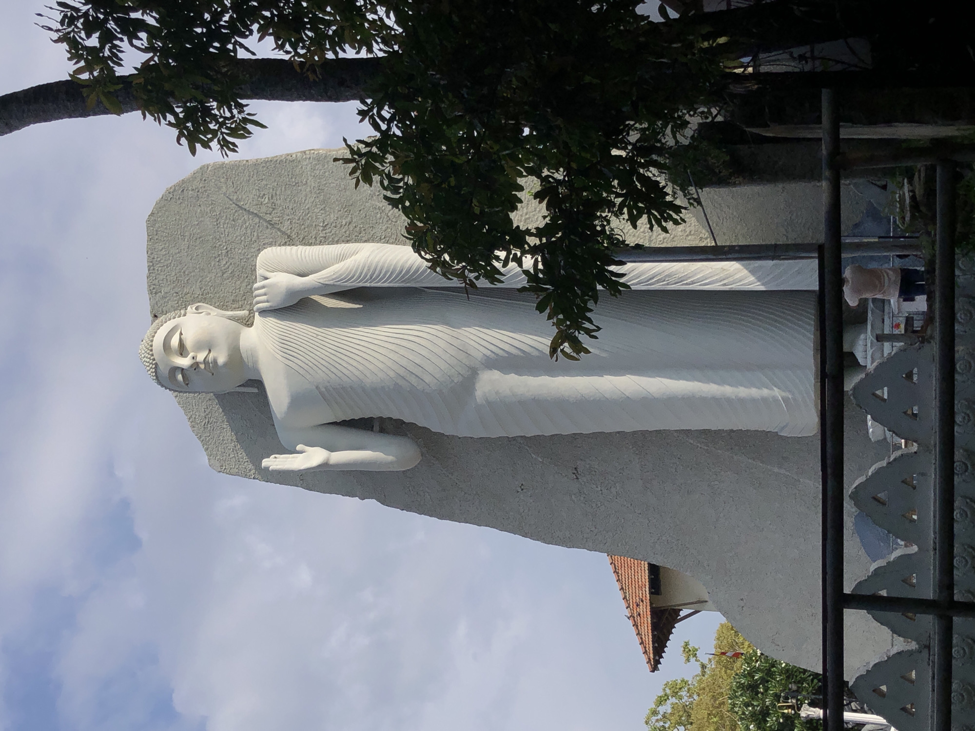 A white Buddha statue lies horizontally, resting on its right side, against a textured grey backdrop. A tree branch with green foliage hangs above the statue. Part of a building with a red-tiled roof is visible behind the statue. A black, ornate fence appears on the right side of the frame. The sky is light blue with clouds. The statue is located in Dondra, Sri Lanka.