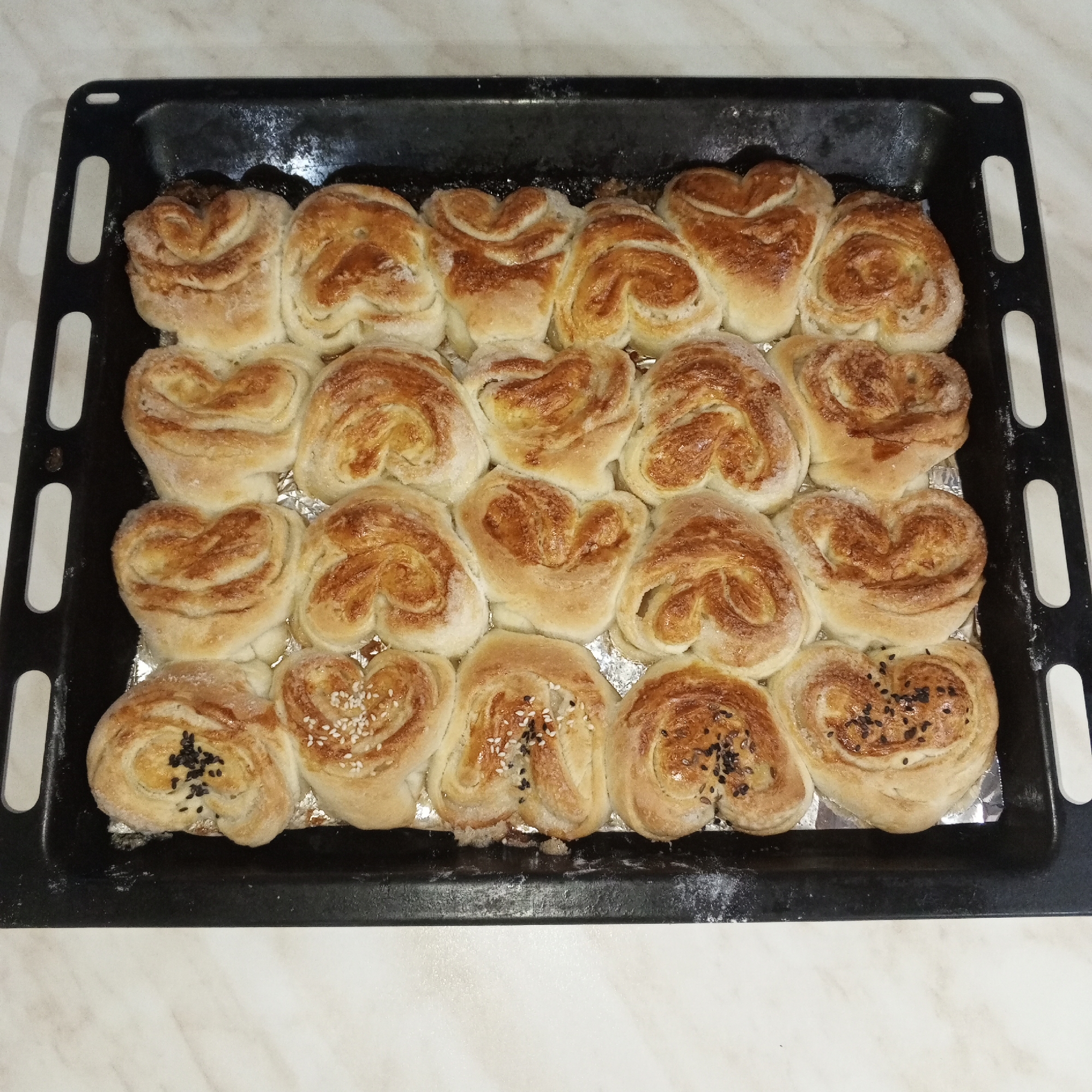 The image shows a black baking tray filled with twenty heart-shaped sweet buns or pastries, arranged in four rows of five. Some buns are sprinkled with white sesame seeds, others with black sesame seeds. The tray appears to be lined with aluminum foil. The buns are golden brown in color, indicating they have been baked. The image is taken in a kitchen setting with a light-colored countertop. The location in the city of Gremyachinsk, Russia, is not visually discernible from the image.