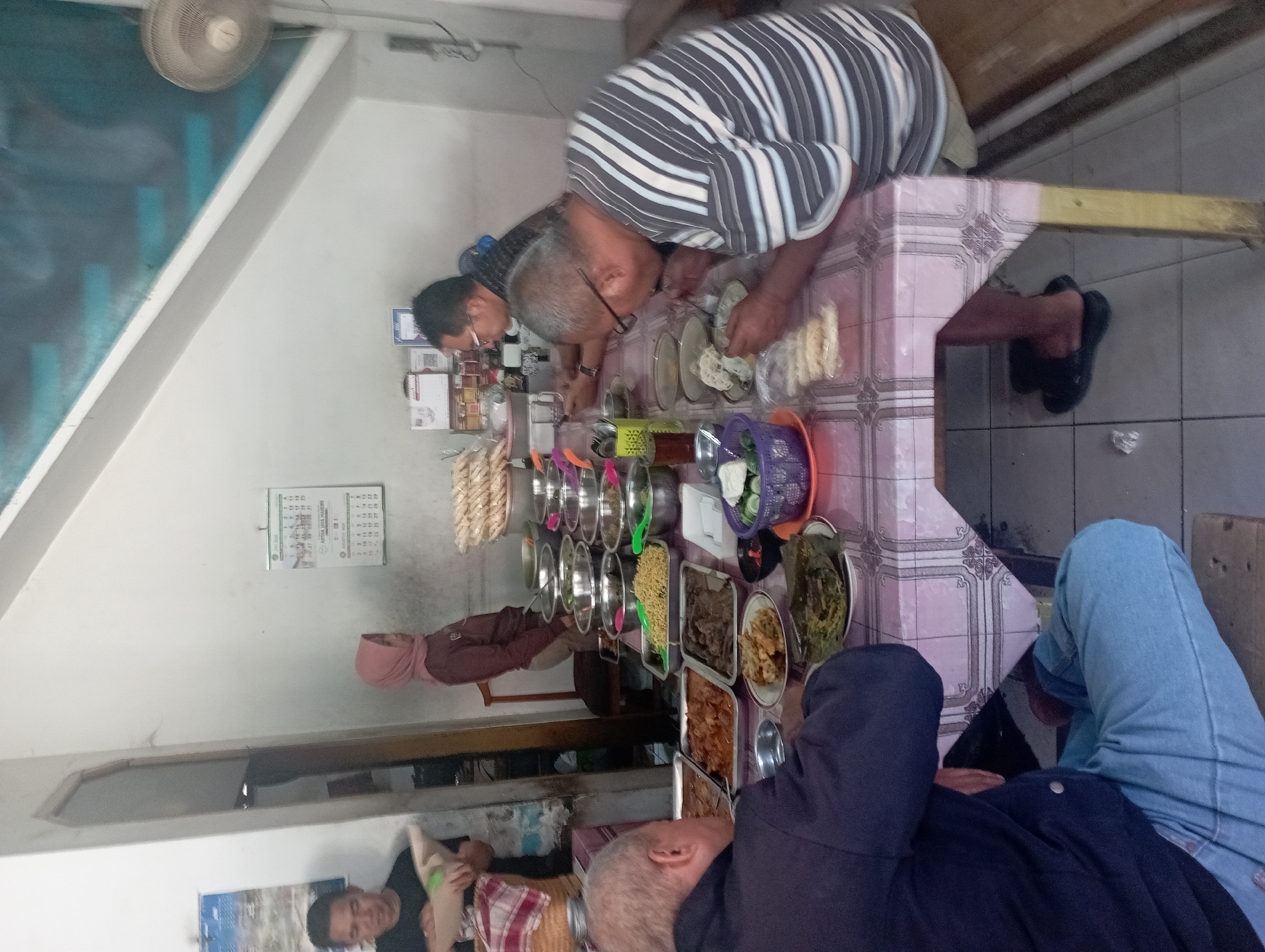 In an indoor establishment resembling a simple eatery, potentially in Tasikmalaya, Indonesia, four individuals are seated around a table laden with various dishes. The table is covered with a pink and white patterned tablecloth. In the foreground, an older man wearing a dark blue jacket and light blue jeans is seated, facing away from the camera, and appears to be eating. To his right, another older man with glasses, dressed in a horizontally striped shirt, is also eating. In the background, a man wearing a dark t-shirt is partially visible, and a woman in a pink headscarf is standing near a doorway. The table displays numerous metal bowls and trays containing food, including what appears to be noodles, meats, vegetables, and rice. Several stacks of crackers are also visible on the table. The room has white walls, and a ceiling fan is mounted on the wall in the background. A calendar is displayed on the wall.
