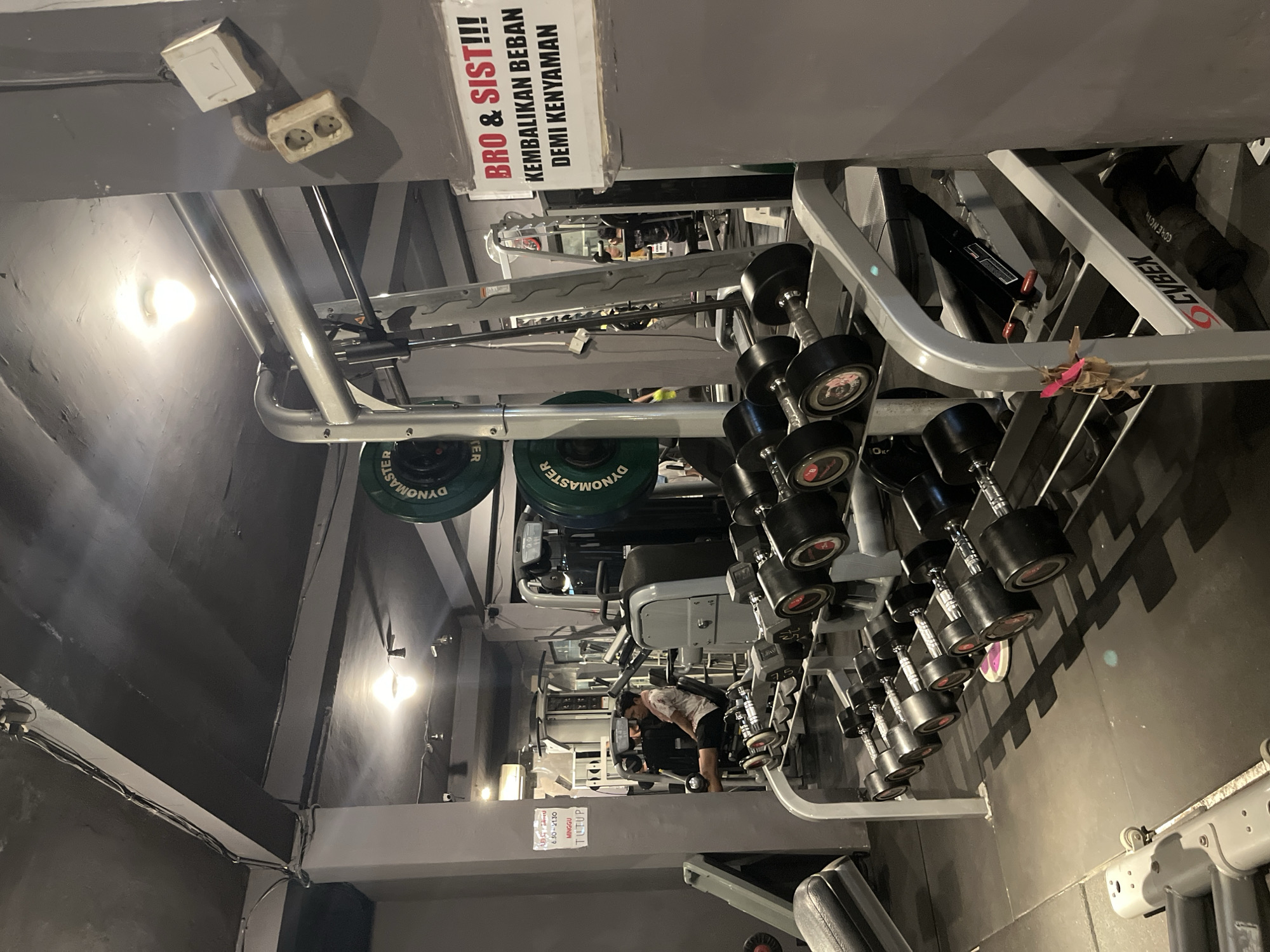 The image depicts the interior of a gym in Denpasar, Indonesia. The scene is dominated by exercise equipment, including rows of dumbbells on racks, weight plates on a rack, and various weight machines. A person is visible in the background using a leg press machine. The gym has a utilitarian aesthetic with exposed ceiling elements, concrete floors, and gray walls. A sign with Indonesian text, "BRO & SIST!!! KEMBALIKAN BEBAN DEMI KENYAMANAN," which translates to "BRO & SIST!!! RETURN THE WEIGHTS FOR COMFORT," is prominently displayed on a gray wall. Electrical outlets and wiring are visible on the wall. The overall impression is that of a functional, albeit somewhat unrefined, fitness facility.