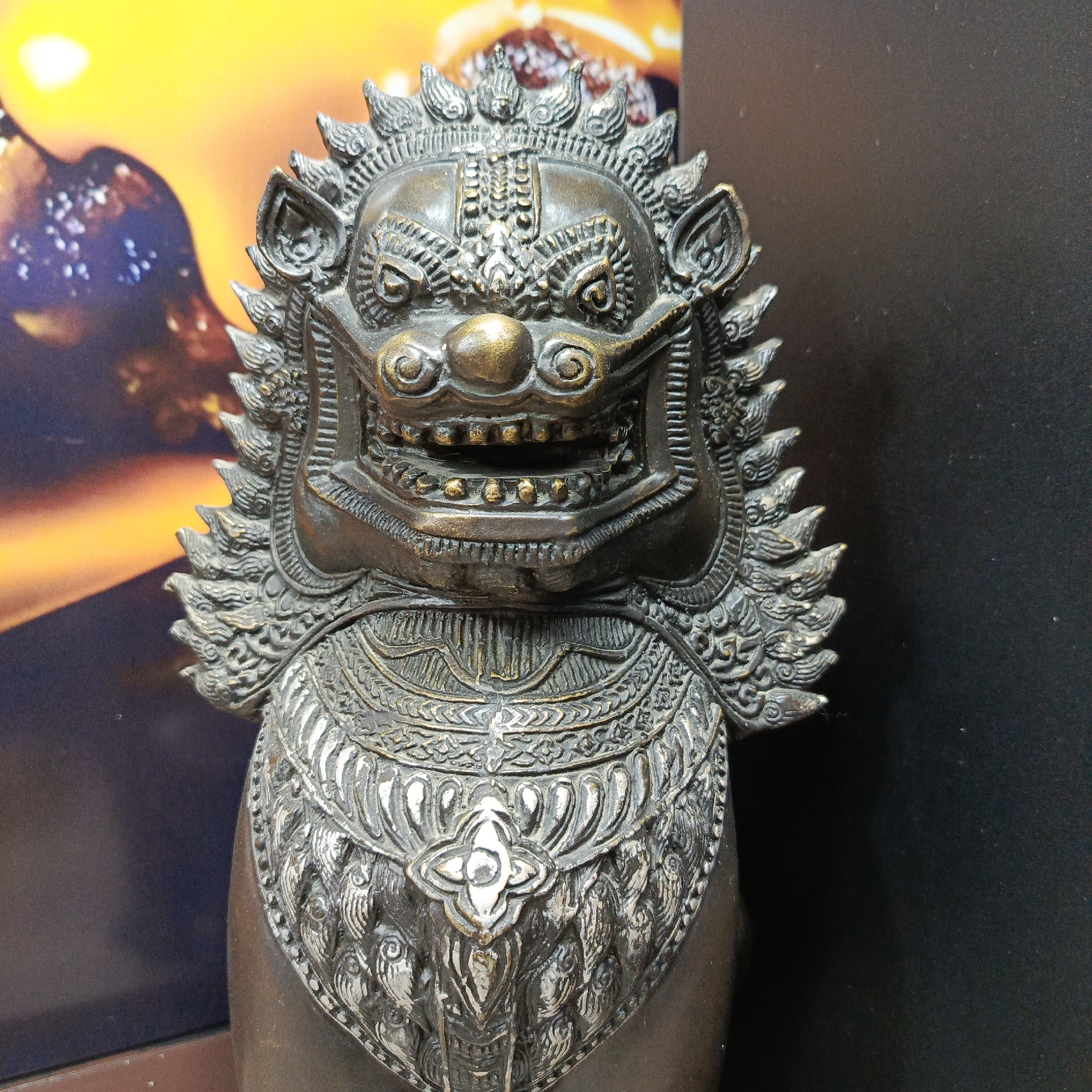 A detailed close-up shot shows a bronze statue of a guardian lion, likely a Foo dog or similar mythological creature. The statue features an ornate collar and intricate designs on its head and mane. The statue appears to be indoors, with a blurred background of what may be an image or artwork, and a solid black background to the right. There is no information in the image to identify the location as Chaikovsky, Russia.