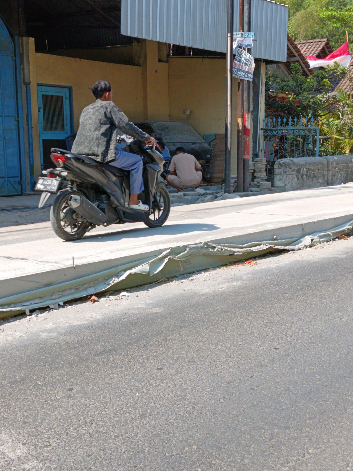 In Barukan, Indonesia, a man wearing a gray patterned jacket and blue pants rides a black motorcycle, its license plate reading "AB 4635 UK". To his right, another man squats by a dark-colored car, possibly working on it. The scene is set on a concrete sidewalk adjacent to a road, with construction materials, including ripped fabric and orange cones, visible at the edge of the sidewalk. Behind the men, a building with a corrugated metal roof stands, with signs attached to a pole in front of it. An Indonesian flag is displayed on a house in the background, and a blue metal fence borders a property. The sun casts shadows, indicating it is daytime.