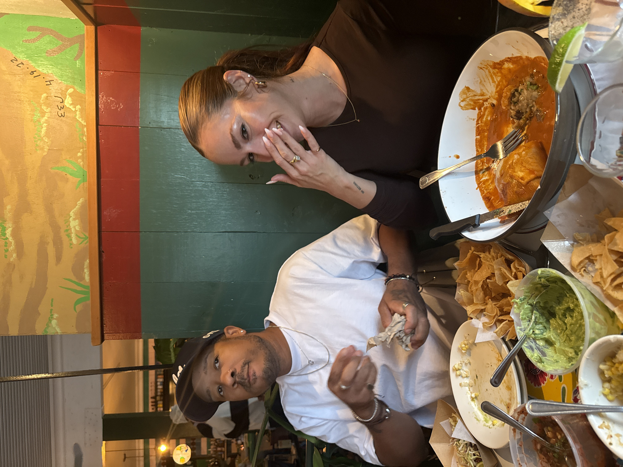 Scene: A restaurant in Los Angeles, United States.
People: A woman with her hair up, wearing a brown long-sleeved shirt, jewelry (necklace, earrings, rings), and a man wearing a white t-shirt, baseball cap, and jewelry (necklaces, bracelets, rings).
Subjects: Plates of food, including enchiladas, guacamole, tortilla chips, salsa, and a dish with corn.
Objects: Silverware (forks, knives, spoons), glassware, containers holding guacamole and salsa, a decorative mural on a wall with green, red, and tan colors.
Actions: The woman is covering her face with her hand, while the man is holding a napkin. Both appear to be at the end of a meal.
Interactions: The pair are seated close to each other at a table covered with food.
Environment: The restaurant has indoor lighting and some decorative elements. A mural on a nearby wall adds color to the setting. Other parts of the restaurant, visible in the background, show additional lighting and a possible bar or service area.
