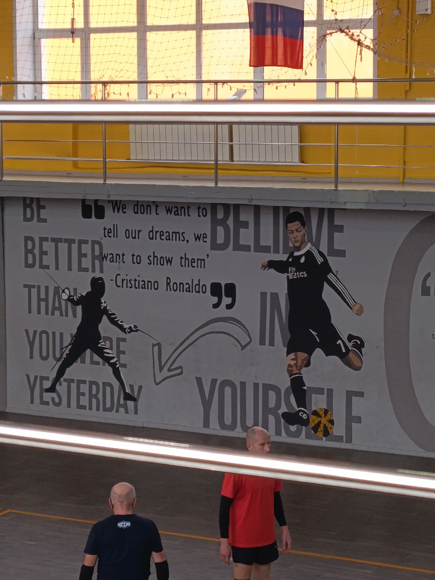The image depicts an indoor sports facility in Krasnoyarsk, Russia. A mural adorns the wall featuring inspirational messages and images of Cristiano Ronaldo, a soccer player, and a fencer. The mural reads, "'We don't want to tell our dreams, we want to show them' - Cristiano Ronaldo BE BETTER THAN YOURSELF, BELIEVE IN YOURSELF." Two men are standing in the foreground: one is wearing a red shirt, black shorts, and black arm sleeves, and the other is wearing a navy blue shirt with 'Россия' (Russia) written on it, and black arm sleeves. A metal railing is visible in the foreground and background. A Russian flag hangs from the ceiling in the background. Windows with netting cover the top section of the background.