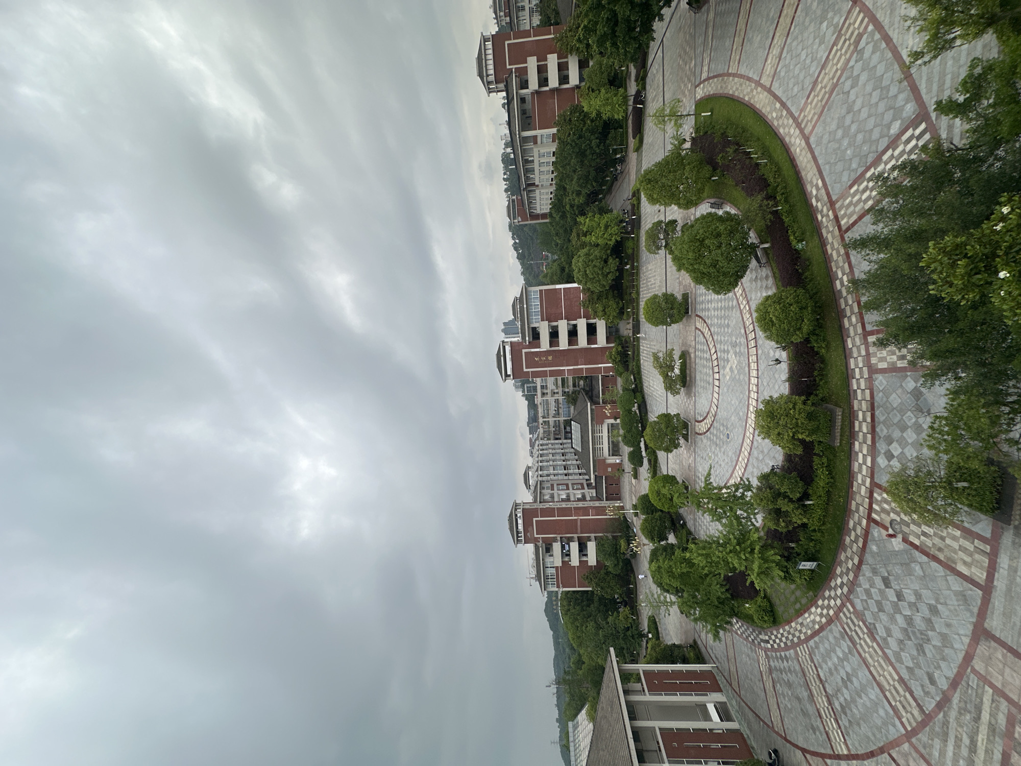 The image is taken from an elevated perspective, overlooking a circular plaza and several buildings, likely a campus setting. The sky is overcast with dense cloud cover.
The plaza is paved with grey and white checkered tiles, patterned in concentric circles, and punctuated by green landscaping including trees and shrubs.
Several multi-story brick buildings with distinct architectural details are arranged around the plaza, possibly dormitories or academic buildings. The surrounding area appears to be hilly with dense vegetation.
The location is identified as Chongqing High-tech Zone, China. There are no people visible in the image.