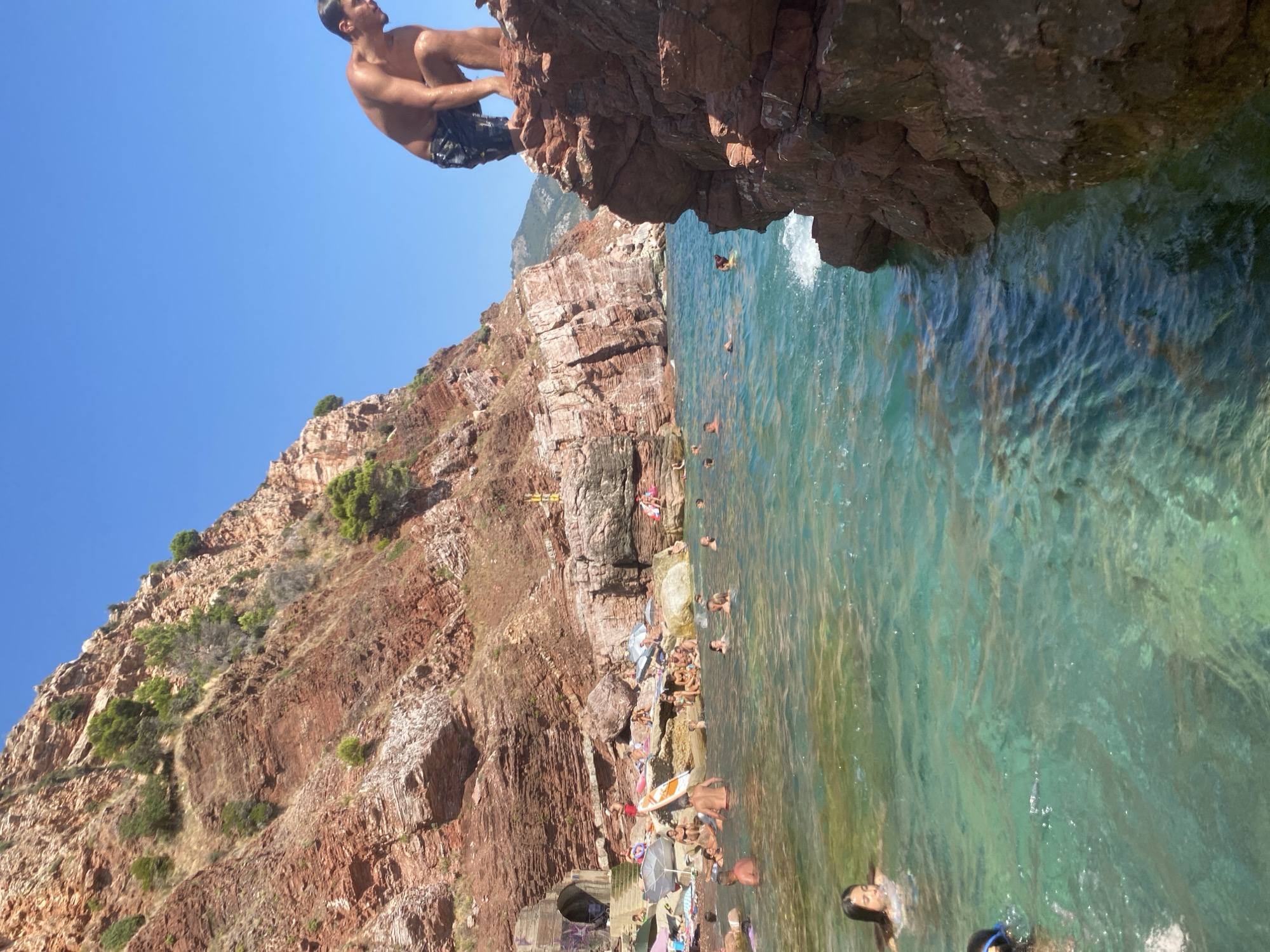 This is an aerial shot of a crowded beach in Sutomore, Montenegro, on a sunny day. The beach is situated at the base of a large, red rock cliff. The water is clear and turquoise, with many people swimming and sunbathing. In the foreground, a man is standing on a rock overlooking the water. In the background, there are more people on the beach and scattered across the water. The cliff face is steep and covered with vegetation. There are also some small caves or openings in the cliff.