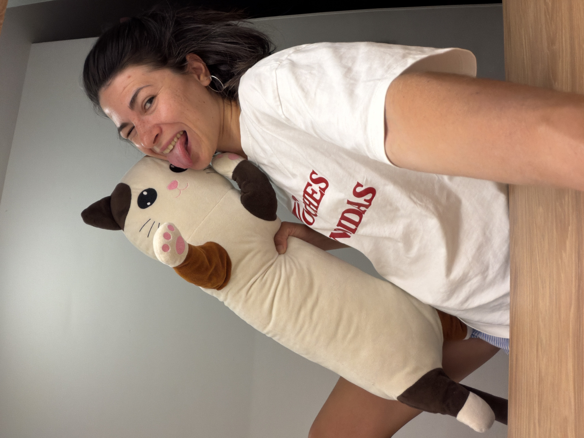 A young woman is holding a stuffed cat in an indoor environment. She is wearing a white t-shirt with red lettering and is winking and sticking out her tongue. The stuffed cat is long and cylindrical, with brown and white coloring. The background consists of a gray wall and a wooden surface. The location is not identifiable as Budva, Montenegro.