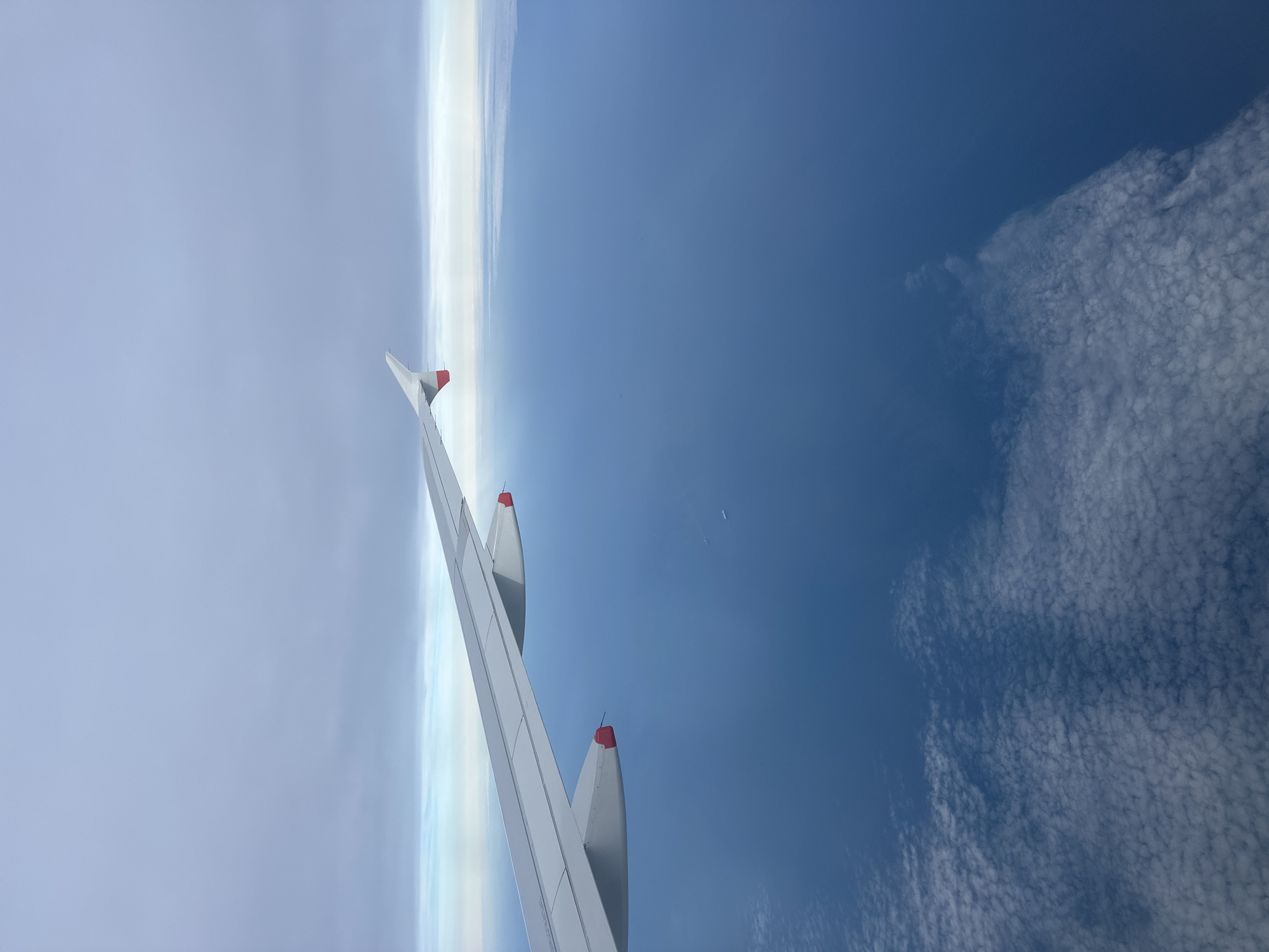 An aerial view from an aircraft window captures a white airplane wing extending from the lower-left to the upper-right of the frame, featuring an upward-curving wingtip with a red marking on its leading edge and two visible engine pylons with red markings underneath. The upper half of the image presents an overcast sky transitioning from a light grey-blue at the top to a brighter, lighter blue and white cloud layer at the distant horizon. The middle and lower-right sections are dominated by a vast, deep blue ocean surface. Two small, white vessels are discernible on the ocean, appearing as minute specks in the mid-right. In the lower-left and bottom-center, a textured layer of white, rippled clouds (likely stratocumulus or altocumulus) partially obscures the ocean directly below the aircraft. The scene is illuminated by diffuse daylight.