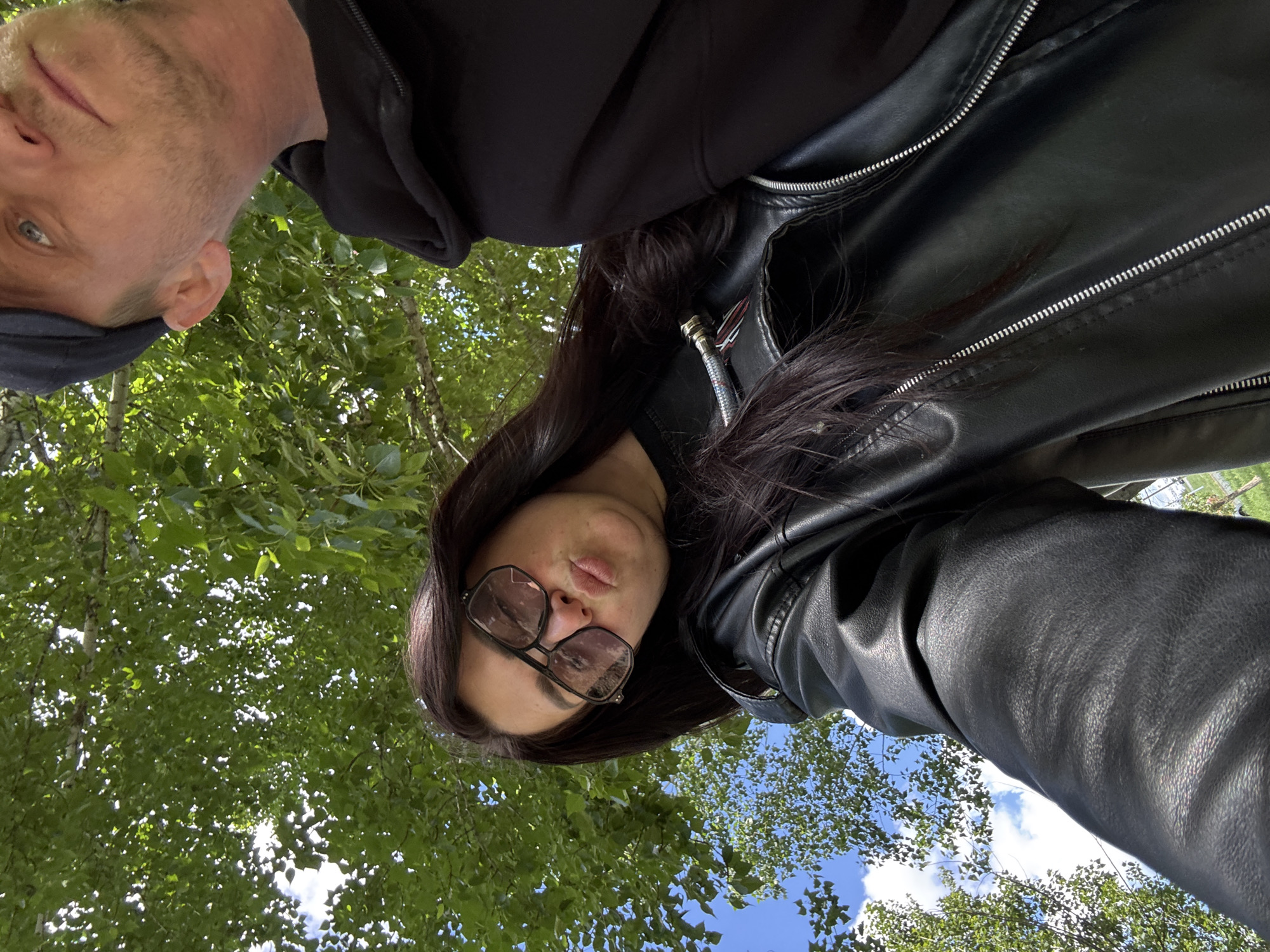 An upward-angled outdoor selfie captures two individuals against a backdrop of vibrant green tree foliage and a bright blue sky with scattered white clouds.
On the left side of the frame, partially visible, is a male with short, light-colored hair and some facial stubble. He wears a dark hooded top or jacket and a dark cap, with his left eye and a portion of his nose and mouth visible as he looks towards the camera.
More prominently featured on the right is a female with long, dark hair. She wears large, dark-framed sunglasses and a black leather jacket with visible silver zippers. Her lips are slightly pouted, suggesting a playful or stylized expression.
The overall perspective of the image is from a low viewpoint, looking up at the subjects, giving the impression that the photo was taken from below them. The ample sunlight illuminating the leaves and parts of the sky indicates it is daytime. No text is visible in the image.