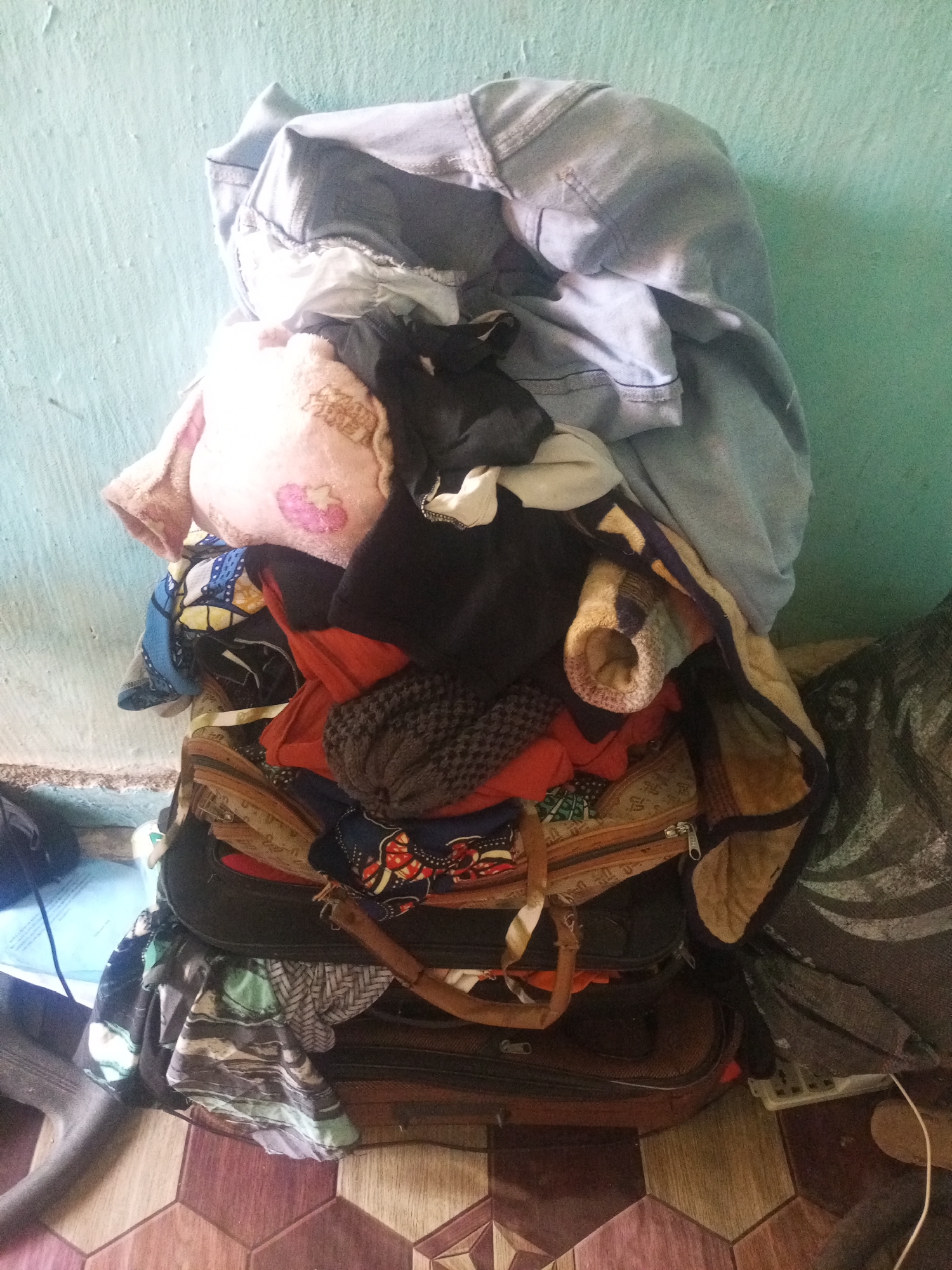 The image depicts a pile of clothes and luggage leaning against a light blue-green wall. The clothing is a mix of colors and textures, including what appears to be a light blue denim jacket or shirt at the top, a pink plush item with embroidery, and various other garments in shades of black, red, orange, and patterned fabrics. Beneath the clothes, several pieces of luggage are stacked. The luggage appears to be dark colored, with zippers and handles visible. One piece of luggage has a brown leather strap. The floor beneath the pile is tiled with hexagonal tiles in shades of brown and wood grain patterns. To the right of the pile, a portion of a dark gray bag with white lettering is visible, leaning against the wall. The scene is consistent with a domestic environment or storage area, potentially in Jalingo, Nigeria, given the context provided. There are no people visible in the image. The lighting suggests an indoor setting.
