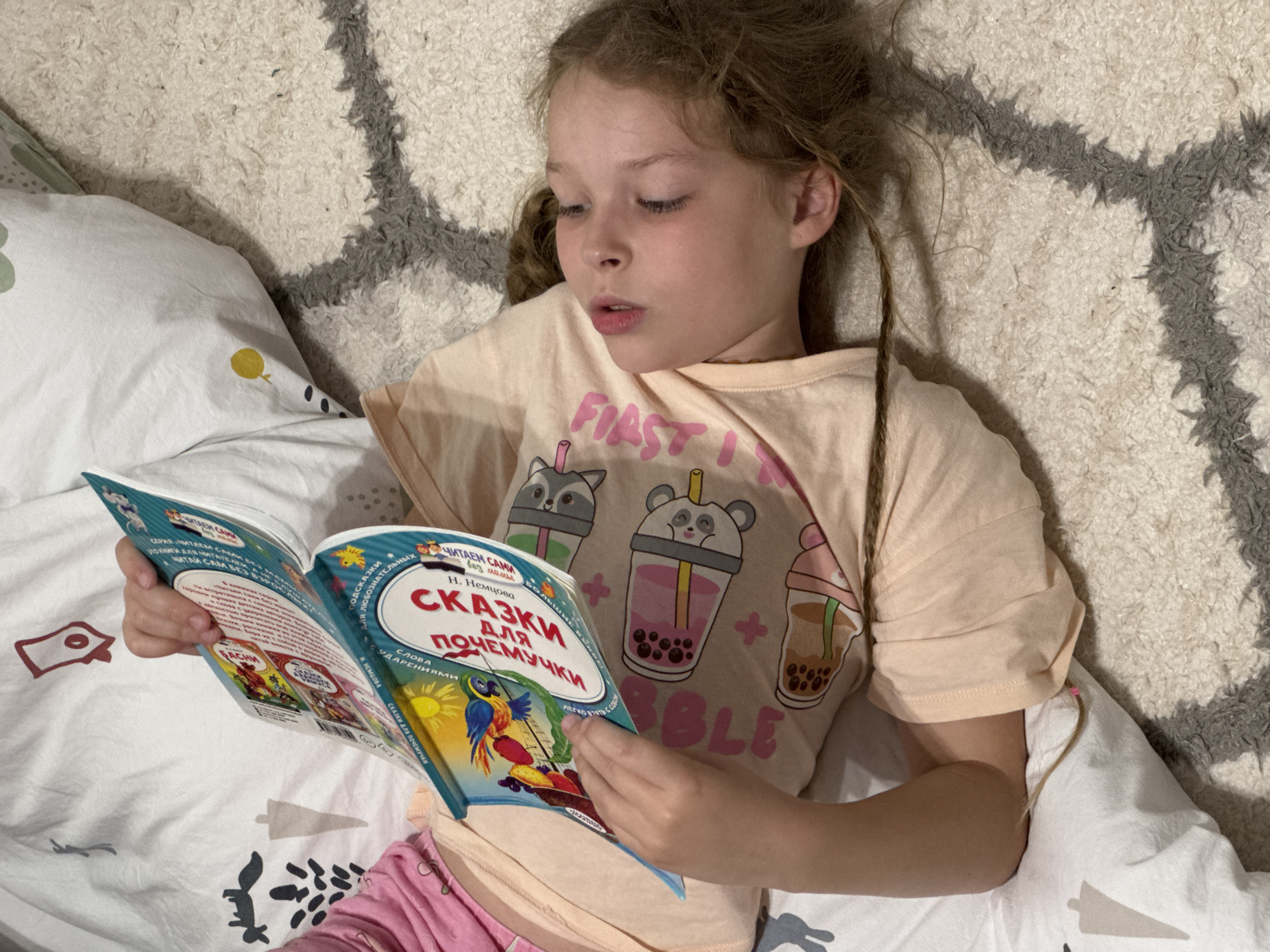 A young girl with light brown, curly hair is lying down indoors, absorbed in reading a book. She wears a light peach-colored short-sleeved t-shirt with cartoon illustrations of bubble tea cups featuring a raccoon, panda, and another animal. Pink text on her shirt, partially visible, reads "FIAST" and "BBLE". She also wears pink pajamas.
She holds an open book with a blue cover. The prominent title on the right page is "СКАЗКИ для ПОЧЕМУЧКИ" (Tales for a Curious Child), with the author "Н. Немцова" (N. Nemtsova) and an illustration of a colorful parrot. Text on the left page includes "ЧИТАЕМ САМ" (Read Myself) and phrases like "СЕРИЯ ЧИТАЕМ САМИ БЕЗ МАМЫ" (Series: Read Ourselves Without Mom) and "ЧИТАЙ САМ БЕЗ ВЗРОСЛЫХ" (Read Yourself Without Adults), indicating it's a book for independent young readers. The girl's eyes are focused on the pages, and her mouth is slightly open in an expression of deep concentration.
She is lying on a white textured blanket or duvet, partially covering a white pillow with a yellow circle and black dots. The background consists of a shaggy white rug with a distinct gray geometric pattern. The scene depicts a calm and cozy moment of a child reading, likely in a bedroom or living room in Budva, Montenegro.
