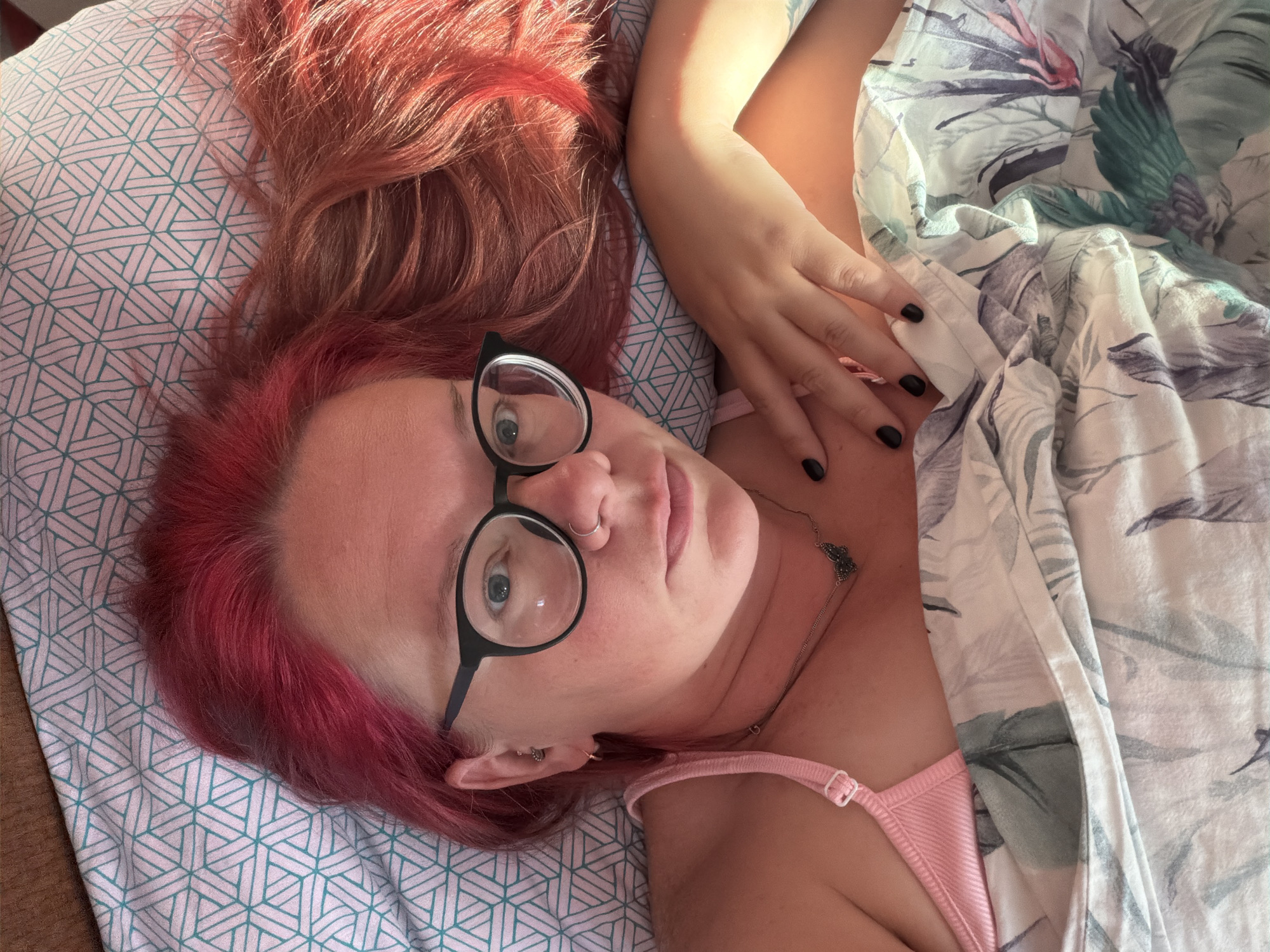 A close-up, high-angle, overhead shot captures a person lying in bed, with their head resting on a pillow covered by a white and teal geometric pattern. The person has bright pink hair, wears black-rimmed glasses and a small silver nose ring. They are wearing a pink camisole and a silver necklace with a butterfly pendant. Their left hand rests on their chest, fingers curled, with black nail polish visible. The person is partially covered by a comforter with a tropical floral pattern featuring large leaves in shades of green, blue, and pink. The image's lighting suggests it was taken indoors, possibly during the daytime, with sunlight illuminating the right side of the person's face and hair. The scene is devoid of any recognizable urban or natural landmarks that would indicate a specific city or location within Chaikovsky, Russia.