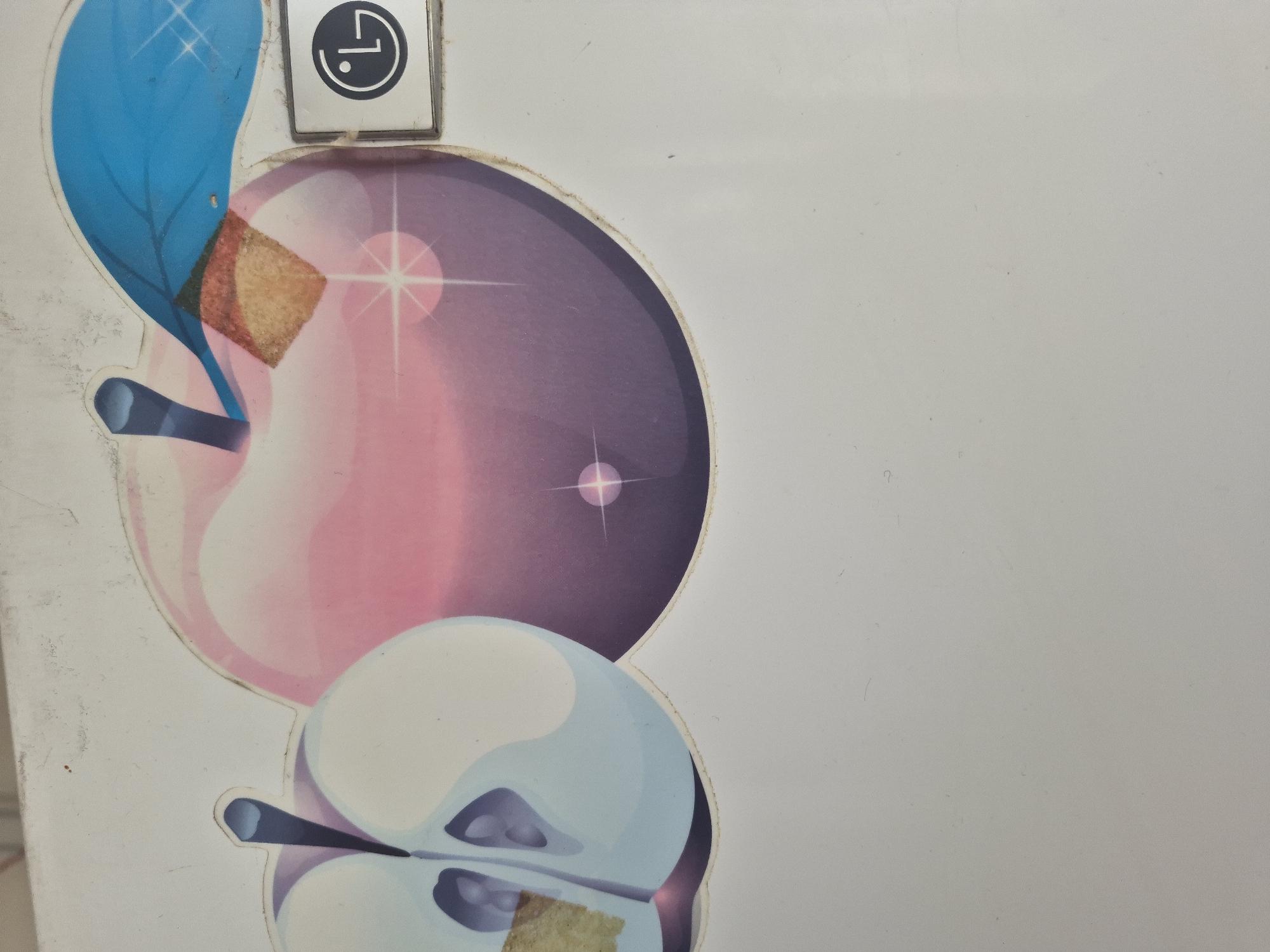 The image shows a close-up view of a refrigerator door with a sticker on it. The sticker features an illustration of two stylized apples and a blue leaf. One apple is depicted whole and is colored in shades of pink and purple with sparkling highlights. The other apple is cut in half, revealing a light blue interior with darker blue seeds. To the right of the apples, there is a rectangular silver and white emblem with a black outline containing the LG logo. The refrigerator door itself is a dull white color and appears to be slightly worn with some minor scuffs and dust. The overall scene is a still life composition focused on the decorative sticker and the LG branding. The location in Yerevan, Armenia, is not discernible from the visual information provided in the image.
