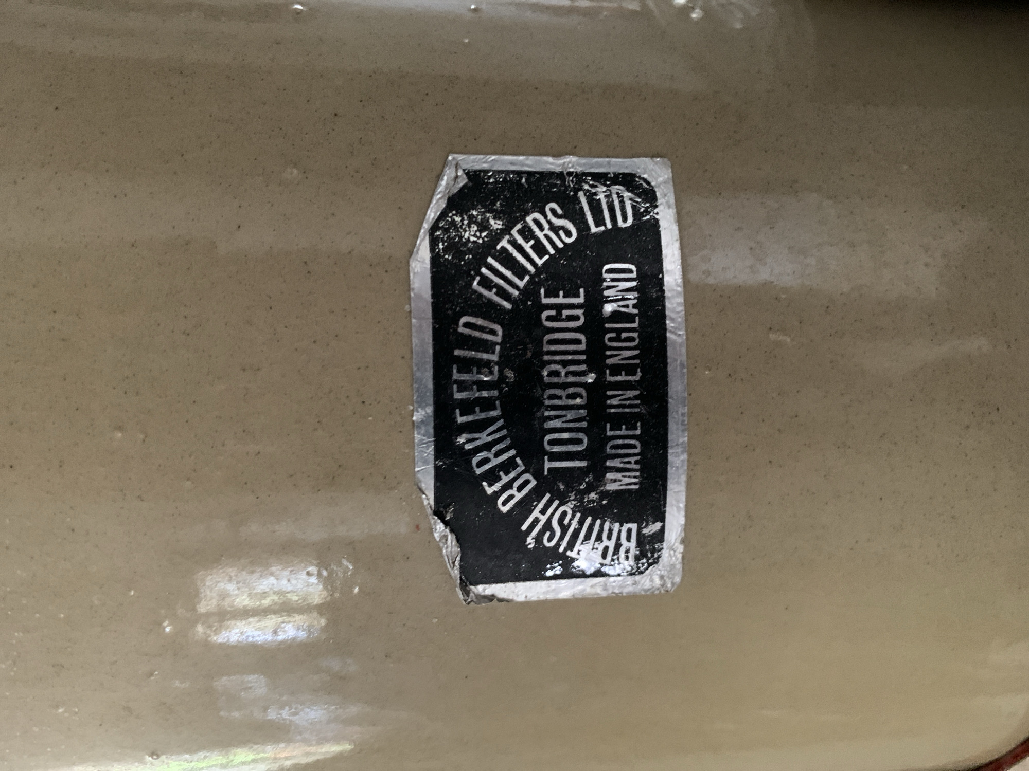 A close-up shot features a distressed black and white label affixed to a textured, light brown or beige surface, possibly ceramic or glazed porcelain. The label is rectangular with slightly rounded corners, but appears curved due to being on a cylindrical or rounded object, and is peeling at the bottom left corner, revealing some silver backing. The text on the label is white, arranged vertically along the curve, and reads: "BRITISH BERKEFELD FILTERS LTD", "TONBRIDGE", and "MADE IN ENGLAND". The lettering for "BRITISH" is partially obscured by the peeling, but "BERKEFELD FILTERS LTD" is clearly legible. The word "TONBRIDGE" is visible below "FILTERS LTD", and "MADE IN ENGLAND" is vertically aligned to its right. The surface around the label shows subtle variations in shade and texture, suggesting a slightly uneven or aged material. There are faint reflections on the surface, indicating ambient light, but no specific time of day or weather is discernible. The image provides a detailed view of the product's origin and brand. This photograph was taken in Santa Ana, Venezuela, showcasing an item manufactured in England.