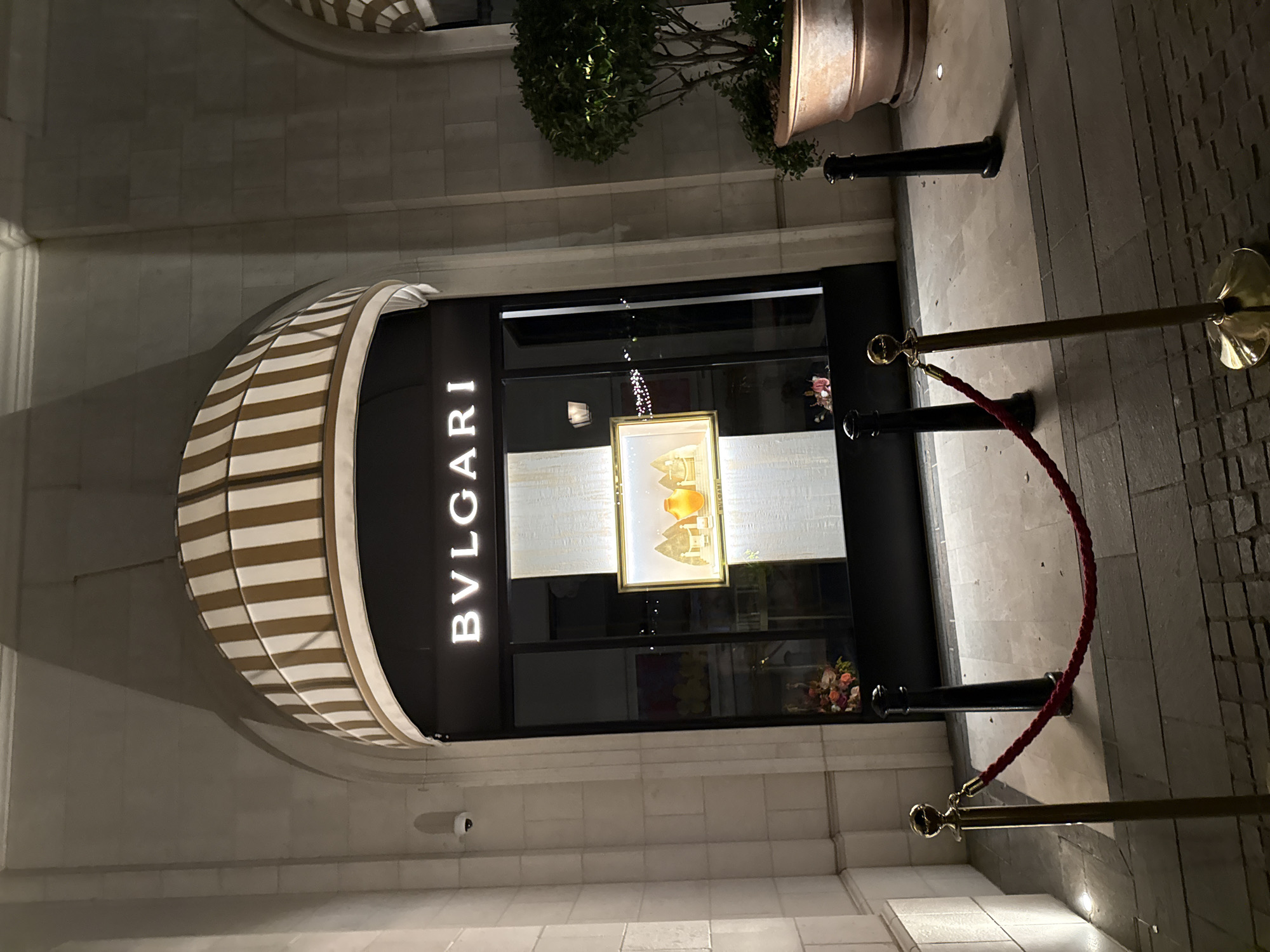 The image depicts the exterior of a Bvlgari store at night, located in Tivat, Montenegro. The store's facade features illuminated "BVLGARI" lettering above a large display window. Inside the window, a framed display showcases decorative bottles against a textured background. A bouquet of flowers is visible on the left side of the window. Red ropes supported by brass stanchions are cordoning off the entrance. A large planter with a green shrub is situated to the right of the store, and a security camera is mounted on the left wall. The ground is paved with stone tiles.