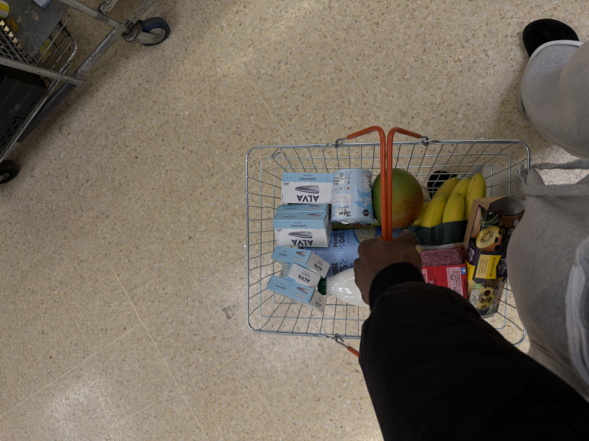 An overhead shot captures a person's arm and leg as they hold a wire shopping basket filled with groceries, likely inside a supermarket in London, United Kingdom. On the left, part of a metal shopping trolley is visible. The floor is covered in light-colored terrazzo-style tiles.
The shopping basket contains a variety of items. Prominently stacked are several light blue boxes of "ALVA SARDINES in Brine". Next to these is a can, partially obscured, with visible text including "PURE CREME coconut". A green and red mango, a bunch of yellow bananas, and a package of "J Sainsbury's PORRIDGE OATS" are also in the basket. Other items include a clear plastic container of "GOLDEN KIWI" with text "Intensely juicy with a sweet tropical flavour," "20p off," and "500g." A package of what appears to be ground meat, marked "PR" and "500g," and a white bottle are also present. The person's arm, wearing a black long-sleeved top with a ribbed cuff, holds the orange handle of the basket. The person's leg, clad in light grey sweatpants, is visible to the right. The presence of "J Sainsbury's" branding confirms the location as a Sainsbury's supermarket.