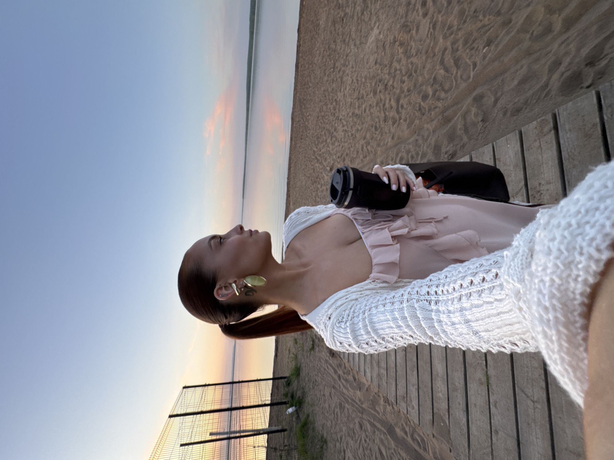 A young woman is at a beach. She has fair skin, brown hair in a low ponytail, and is wearing large, round, gold earrings. She is wearing a pink, ruffled top, and a white, knit sweater. She is holding a black travel mug. She is standing on a wooden boardwalk, with a sandy beach to her right. In the background, there is water meeting a sandy shoreline. To her left, there is a metal fence with a view of the sky at sunset. This is in Chaikovsky, Russia.