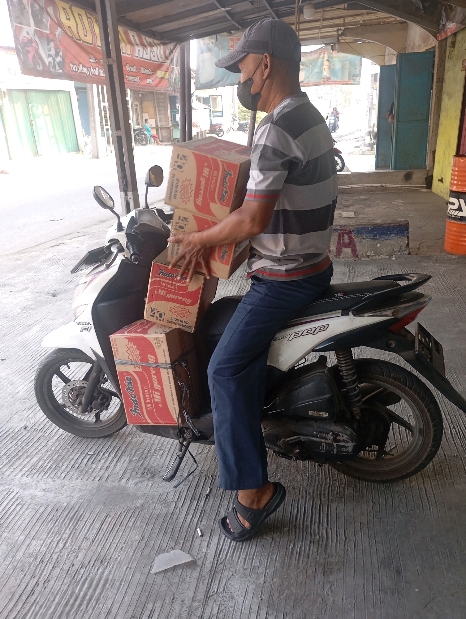 A man wearing a grey and black striped shirt, dark blue pants, a black face mask, and a grey baseball cap is sitting on a white Honda Beat motorcycle. He is holding two stacks of Indomie Mi Goreng noodle boxes. The boxes are red and white. The motorcycle is parked on a textured concrete surface. In the background, there is a covered market or shop area with banners and doorways. The scene appears to be in Babelan, Indonesia, based on the presence of the brand "Indomie" which is popular in Indonesia and common in the region, and typical street architecture and signage visible in the background.