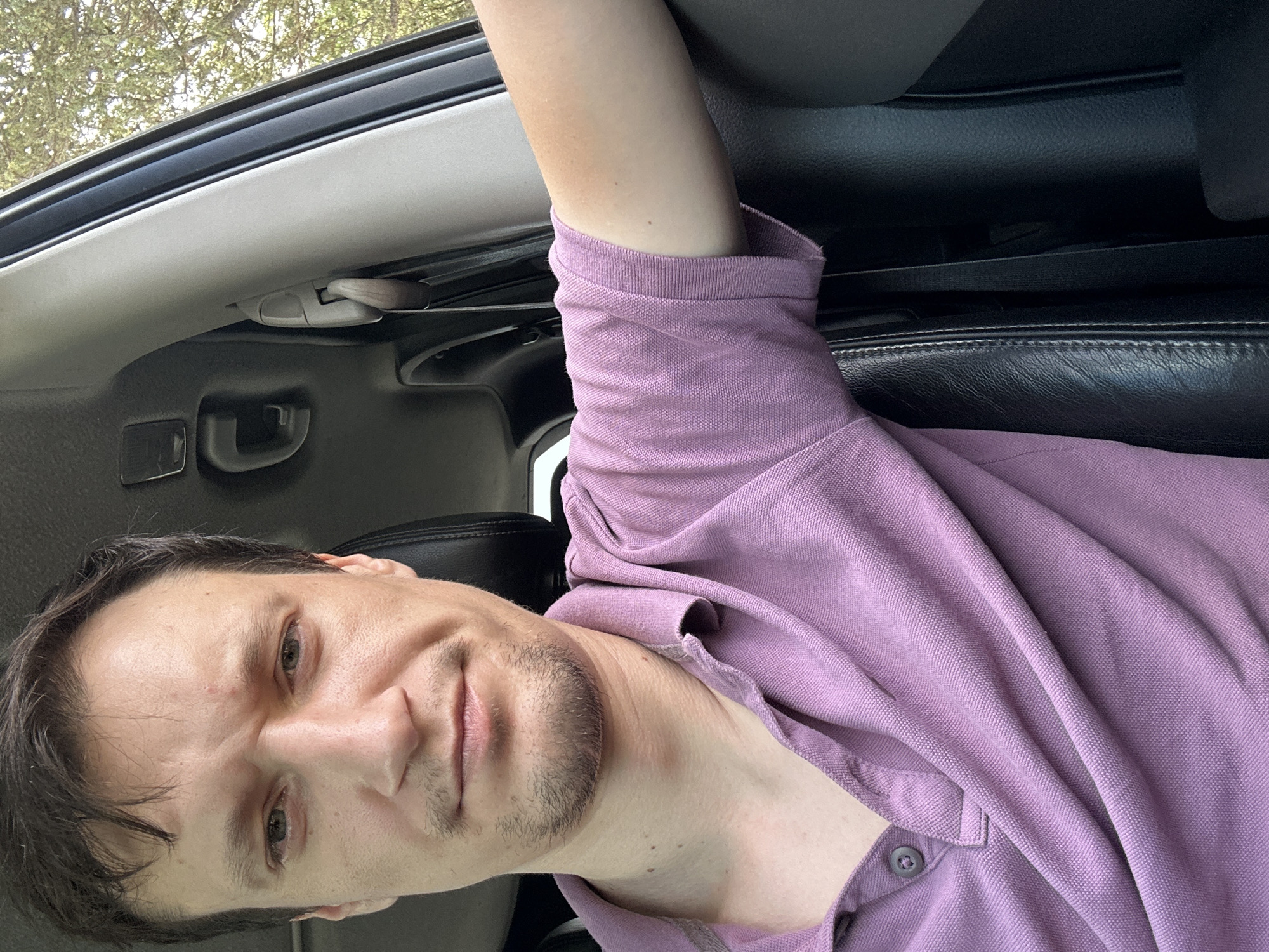 A man with short brown hair and a light complexion is seen from the chest up, taking a selfie inside a car. He is wearing a short-sleeved, light purple polo shirt with a collar and a single button at the neckline. His left arm is resting on the doorframe, and the interior of the car, including black leather seating and a grey pillar, is visible. Sunlight streams in from the right, illuminating the man's face and arm. Outside the car window, trees and foliage are visible, suggesting an outdoor setting possibly in or near Makiv, Ukraine.