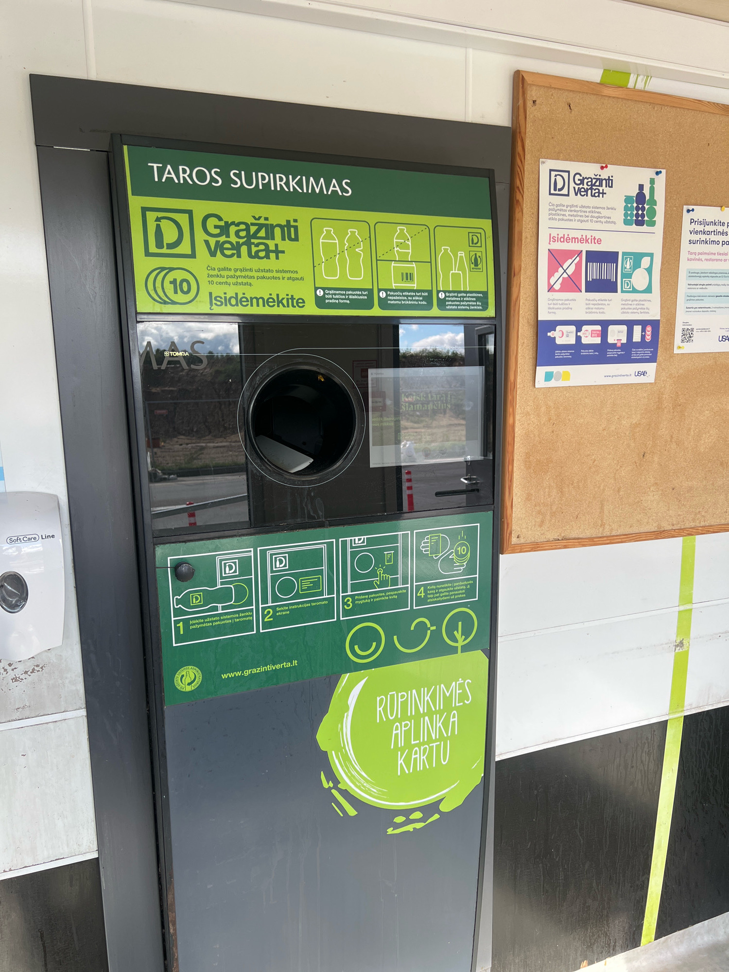 The image shows a deposit return machine for beverage containers, likely located outdoors in a public area in Vilnius, Lithuania. The machine is predominantly black and green, with prominent yellow and white signage. The top of the machine displays "TAROS SUPIRKIMAS" (Container Purchasing) and the "Gražinti verta+" (It's worth returning) logo, indicating its function. There are instructional graphics illustrating how to use the machine, including steps for inserting containers and receiving a voucher. A screen on the machine displays a reflection of the surrounding environment, which appears to be a construction site with dirt mounds and scaffolding. The machine also features a slot for inserting containers and a dispenser for vouchers. To the right of the machine, a bulletin board with informational posters is attached to a wall. The overall environment suggests an urban or semi-urban setting.