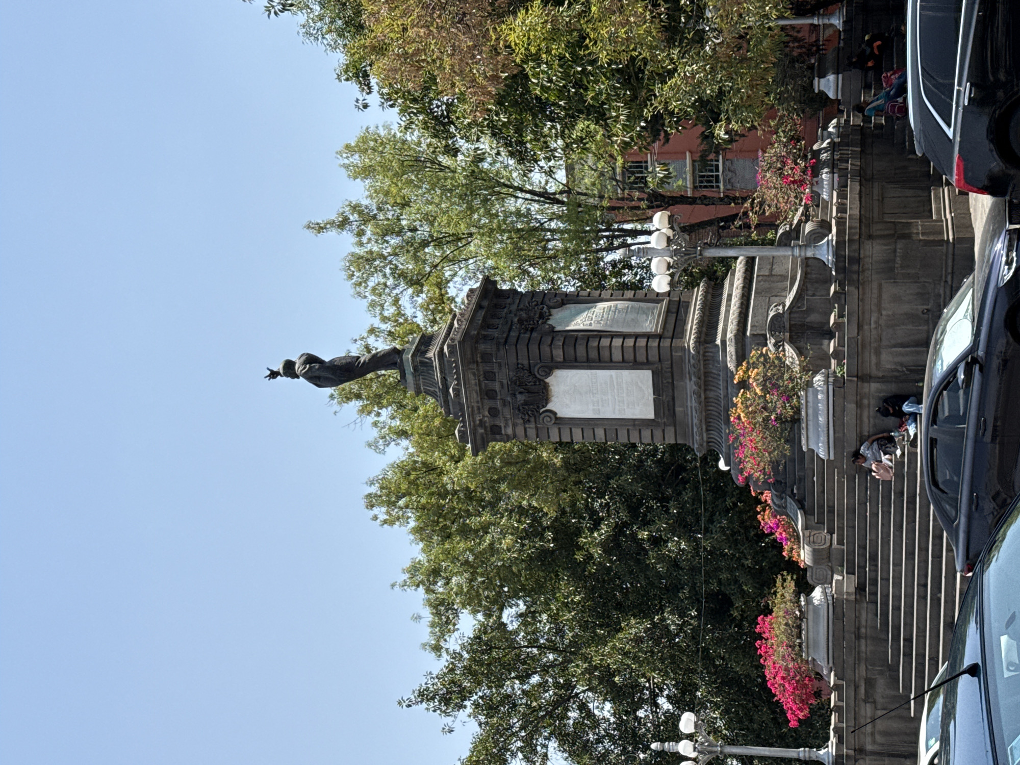 The image captures a sunlit urban scene centered around a tall, ornate monument. Atop the monument stands a statue of a figure, possibly male, with one arm extended, set against a clear, light blue sky. The base of the monument is made of stone, featuring multiple engraved plaques with text, though the specific wording is illegible.
Surrounding the monument are lush green trees, providing shade and contrast to the clear sky. To the right of the monument, a grand stone staircase leads upwards, adorned with decorative planters overflowing with vibrant pink, red, and orange flowers.
Several people are visible in the scene. One individual sits on the lower steps, with a backpack nearby, while another person is further up the stairs, partially obscured. More indistinct figures can be seen in the distance near a reddish building with visible windows, which is partly hidden by the trees.
To the far right, a street is visible with cars. A prominent black car is parked or driving, and parts of another vehicle are seen in the immediate foreground. A traditional street lamp with multiple globes stands near the steps.
The scene depicts a clear, bright day, likely morning or afternoon, with strong sunlight casting shadows. The overall atmosphere is calm, suggesting a public square or park in a city. Given the location context of Puebla, Mexico, this monument is likely a significant landmark within the city.