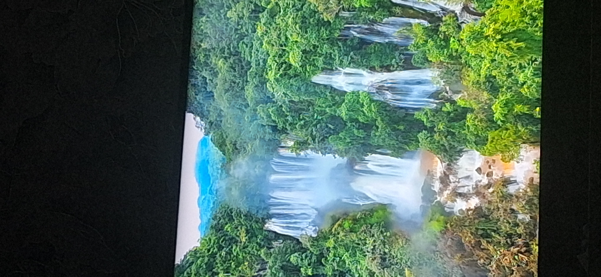 The image displays a breathtaking, multi-tiered waterfall cascading down a lush, jungle-covered cliffside. The water is a milky white, creating a misty atmosphere around the falls. The surrounding vegetation is a vibrant green, with dense foliage framing the powerful flow of water. In the distance, hazy blue mountains are visible, suggesting a vast and natural landscape. The scene is likely a natural wonder, possibly in a tropical or subtropical region, characterized by its untouched beauty and abundant greenery. The image is taken from an elevated perspective, allowing for a panoramic view of the waterfall and its verdant surroundings. There are no people or urban elements present in the scene, emphasizing the wild and remote nature of the location. The provided crops confirm the details of the waterfall, the dense vegetation, and the distant mountains. The image does not depict any part of the city of Bila Tserkva, Ukraine.
