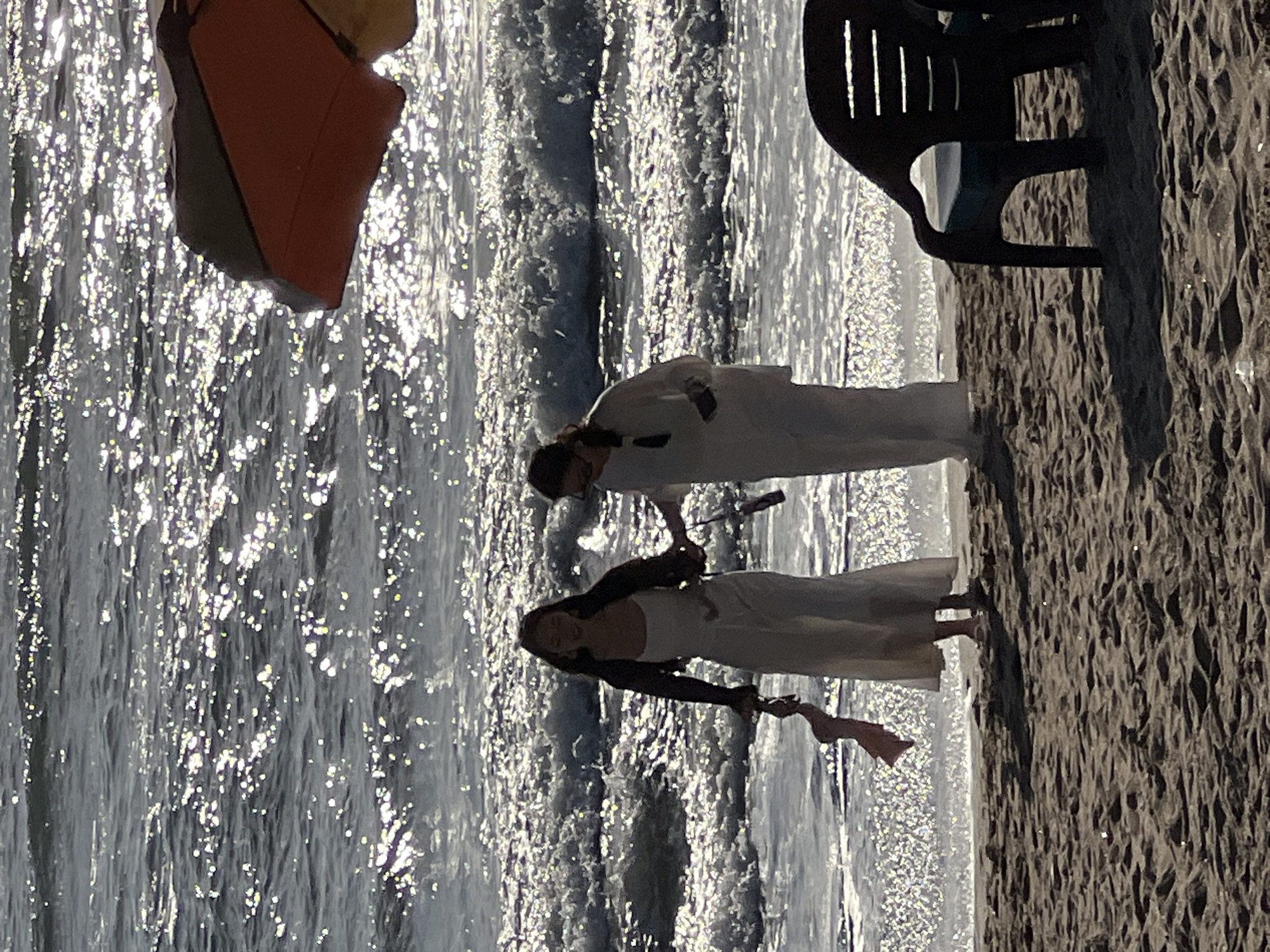 Two figures, silhouetted against the shimmering ocean and sand, walk along the shoreline of Kuta, Indonesia. The figure on the left, wearing a light-colored dress and a dark jacket, holds a pink garment. The figure on the right, also in light-colored attire, appears to be holding an object. Waves gently break on the beach, with sunlight glinting off the water's surface. A dark plastic beach chair is visible in the foreground to the right, and a portion of an orange and yellow umbrella is present at the top right of the frame. The scene is bathed in the warm glow of sunlight.