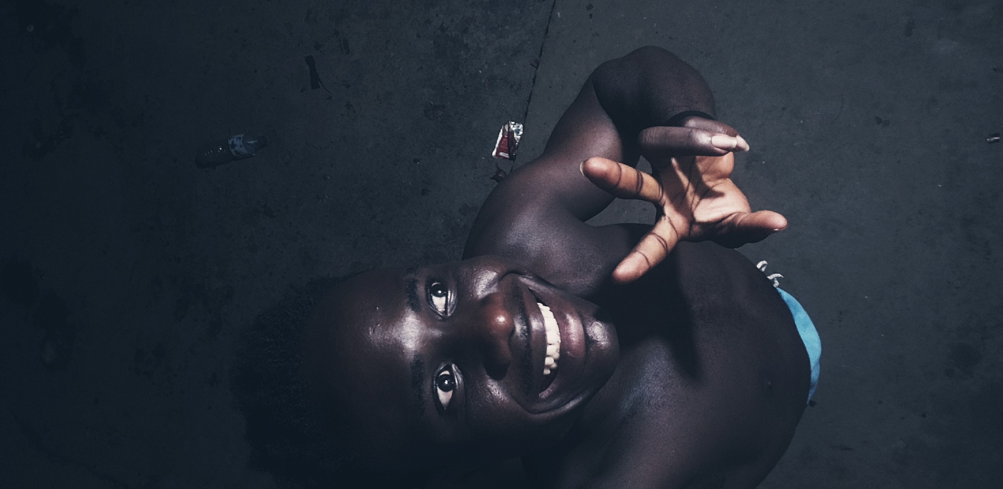 In Narayi, Nigeria, a dark-skinned man is photographed from a high angle, looking up with wide eyes and a large, open-mouthed smile, revealing white teeth. His head is tilted slightly to his right, and he is looking off to his left. His chest and shoulders are bare. His right hand is raised, with his index and middle fingers extended in a peace sign. He is wearing blue shorts. The background is dark and appears to be an outdoor, possibly urban or industrial area with a rough, textured ground. There are a few scattered objects on the ground, including a partially visible plastic bottle and a crumpled red and white object that resembles a cigarette pack or wrapper. The lighting is dim, suggesting it is nighttime or a poorly lit area.