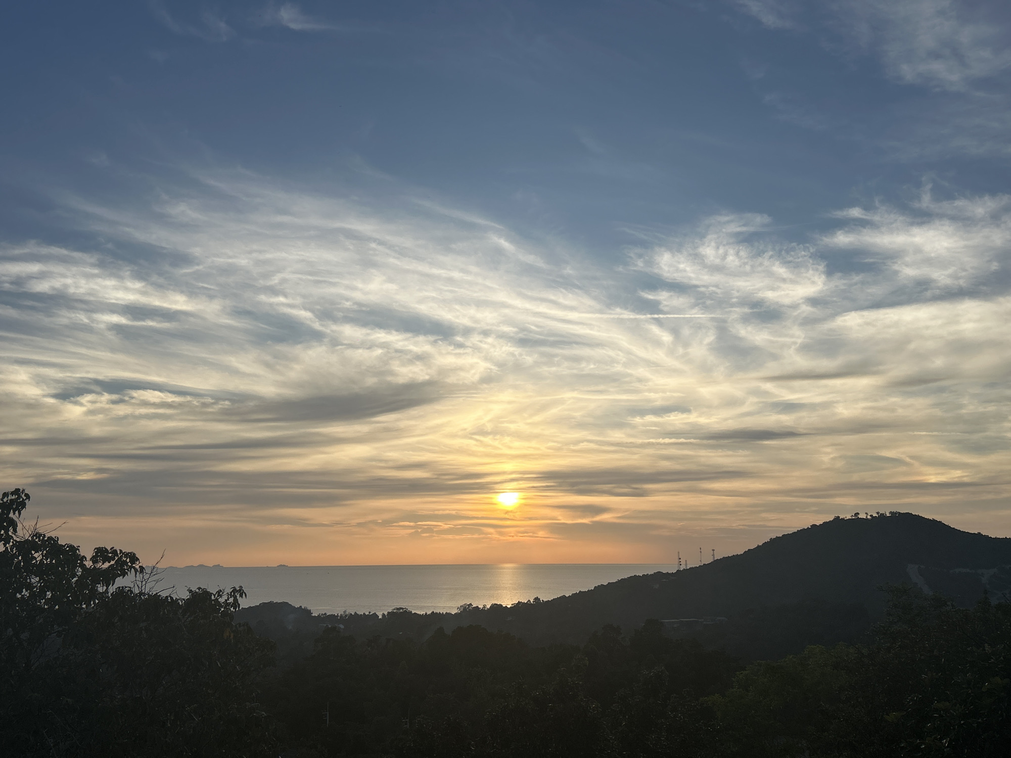 The image depicts a sunset over the sea in ตำบลเกาะพะงัน, Thailand. The sky is filled with wispy, scattered clouds. The sun is near the horizon, casting a golden glow on the water and the lower portion of the sky. A silhouette of a hill with antenna structures can be seen on the right side of the image. The foreground consists of dark, silhouetted trees and foliage. In the distance, small islands are visible on the horizon.