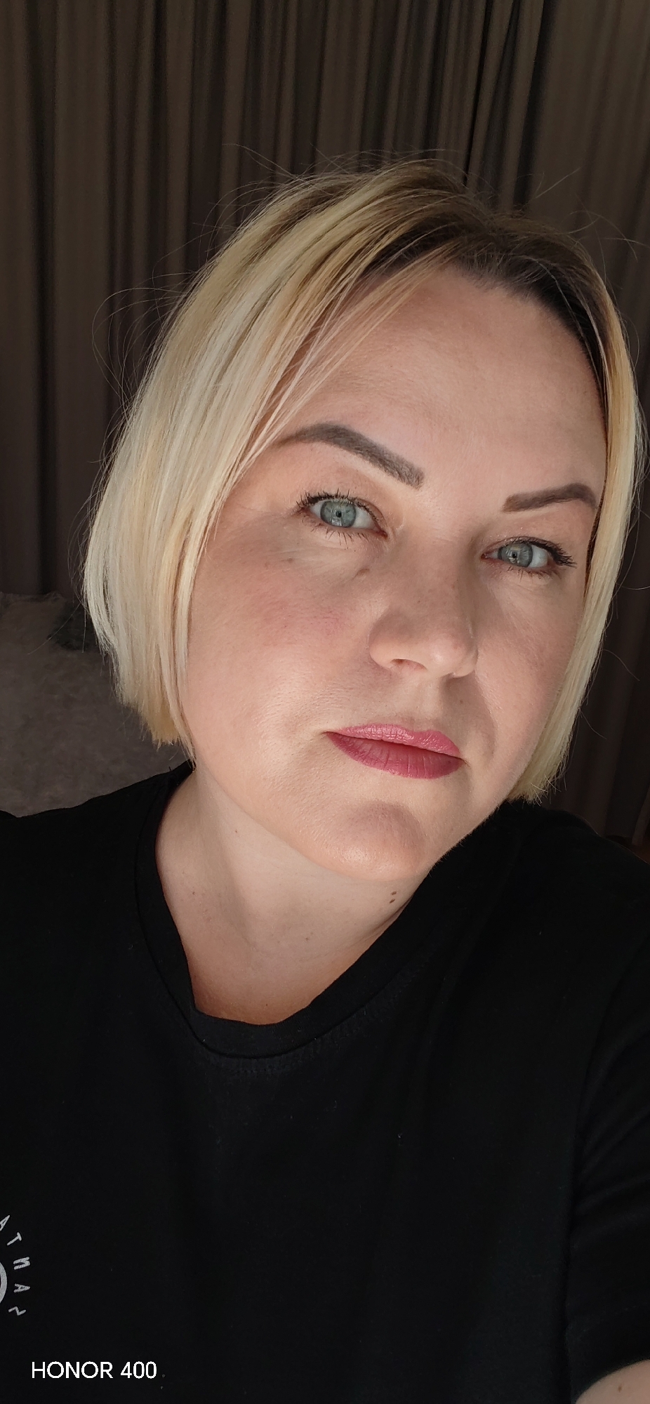 The image depicts a close-up, head-and-shoulders portrait of an adult female in an indoor setting within the city of Chaikovsky, Russia. The subject has short, chin-length blonde hair with visible dark roots, blue-green eyes, and wears pink lipstick. Her expression is neutral, with her gaze directed forward. She is attired in a black crew-neck t-shirt, bearing a partial white circular graphic with the letters "ATMAN" visible on the lower left. The background is blurred, showing dark brown or grey vertical drapery, with a soft, light-colored, textured object partially visible on the left side of the frame. The lighting is soft and appears to be diffused. The image includes a "HONOR 400" watermark in the lower left corner.