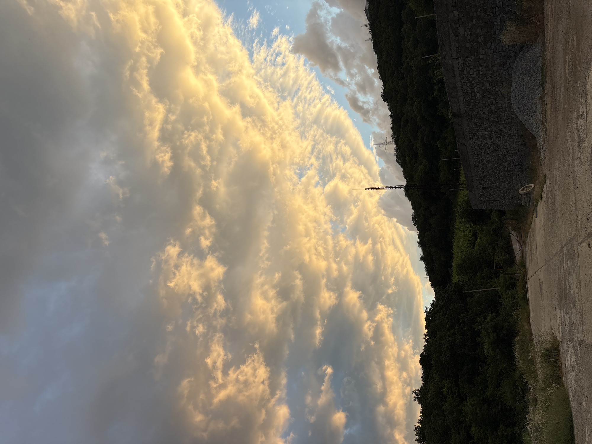 The image depicts a rural landscape under a dramatic, cloud-filled sky, illuminated by the warm hues of sunset. The foreground shows a weathered concrete surface, possibly a road or a courtyard, with patches of dry grass and a small pile of gravel. To the right, a rough stone wall, appearing to be a retaining wall or part of a building foundation, runs horizontally across the frame. Behind the wall, a pile of gray gravel is visible.
Dominating the midground is a densely forested hillside, covered in dark green trees. A faint path or road appears to wind through the trees. Several tall, dark-colored transmission towers, along with what appear to be power lines, are visible on the crest of the hill, indicating the proximity of infrastructure. The sky above is filled with a mix of dark gray and bright yellow-orange clouds, suggesting the presence of both storm clouds and sunset light.
While the location is identified as Malyi Mayak, Ukraine, no specific urban features or inhabitants are discernible in this particular frame, suggesting a remote or undeveloped area. The overall impression is one of natural beauty tinged with the presence of industrial elements.