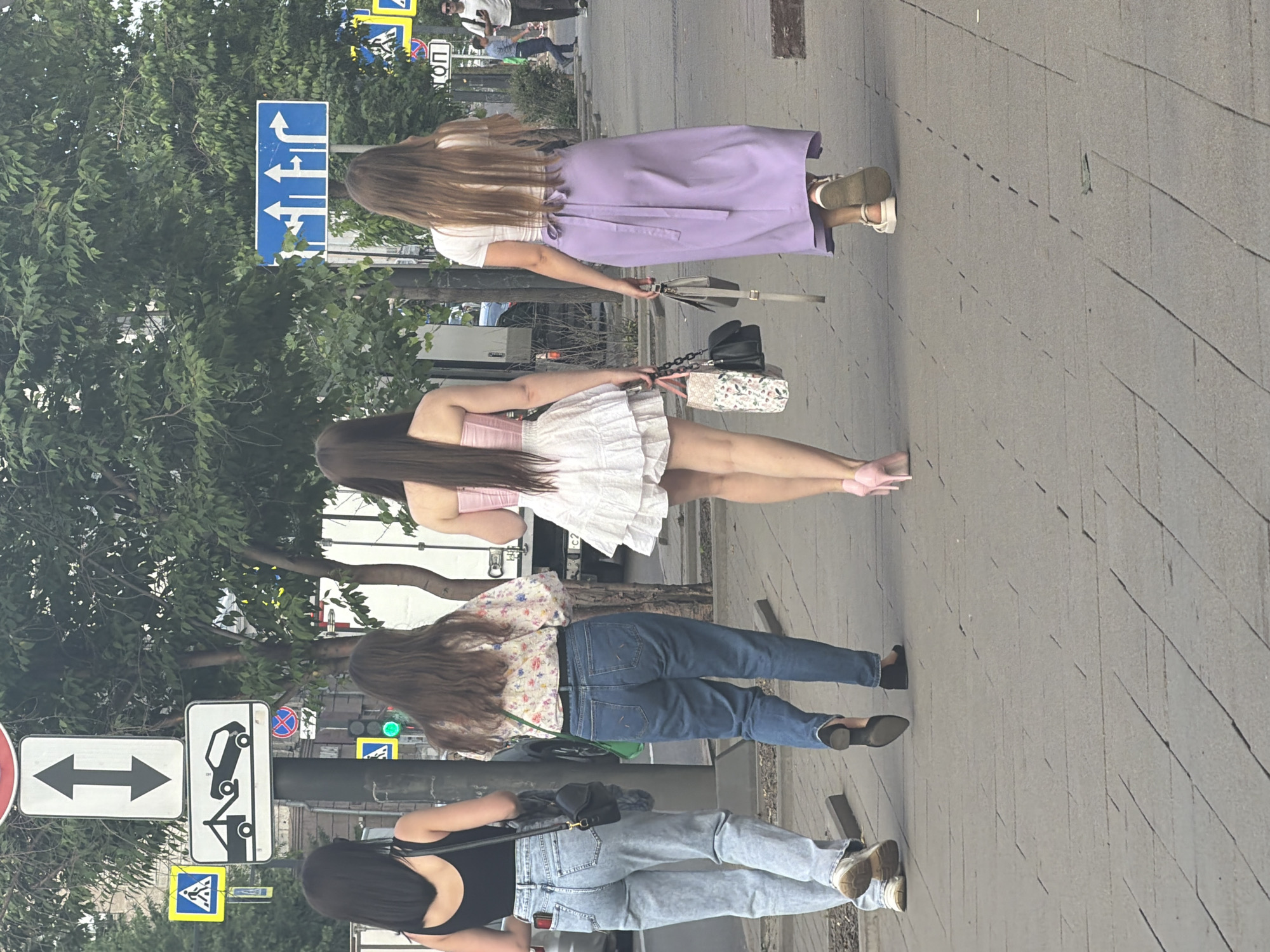 Four individuals, viewed from behind, walk along a paved sidewalk. The person on the far left wears a black tank top and light blue jeans. Next to them, a person in a floral-print blouse and dark blue jeans walks. The third person from the left is dressed in a pink corset-style top and a white ruffled skirt, wearing pink high heels. The person on the far right is in a white t-shirt and a long lavender skirt, with white sandals. Several traffic signs are visible in the background, including a directional sign with multiple arrows, a sign indicating a tow-away zone, and a pedestrian crossing sign. Trees line the sidewalk, and buildings and a white truck are partially visible. The scene is consistent with an urban environment.