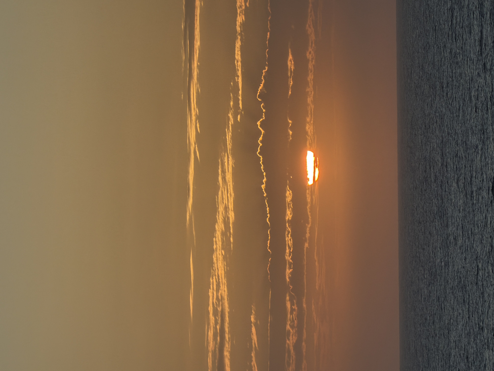 The media file captures a vibrant sunset or sunrise over a large body of water, likely an ocean or sea. The image is oriented vertically, but when viewed horizontally (rotated 90 degrees counter-clockwise), a clear horizon line separates the sky from the water.
The sky, occupying the upper two-thirds of the image, is filled with warm, glowing hues of orange, yellow, and soft brown, indicative of dawn or dusk. Long, thin, streaky clouds stretch across the sky, illuminated by the sun's direct light. These clouds display varying shapes, from relatively smooth lines to more turbulent and wavy formations, casting subtle shadows and highlights against the atmospheric glow.
The sun itself is prominently featured, positioned low on the horizon, partially obscured by clouds or atmospheric haze. It appears as an intensely bright, flattened, and somewhat elongated orange-red shape, radiating strong light that creates brilliant reflections and highlights on the surrounding clouds.
The bottom third of the image is dominated by a vast expanse of water. The water surface is dark, appearing in shades of deep grey or almost black, offering a stark contrast to the brilliant sky. The surface shows a fine texture of ripples or small waves, catching very little of the sun's direct light.
The scene conveys a tranquil yet dramatic mood, characteristic of an atmospheric sunrise or sunset. There are no people, objects other than natural elements, or visible text in the image. The location is an open, unidentifiable seascape.