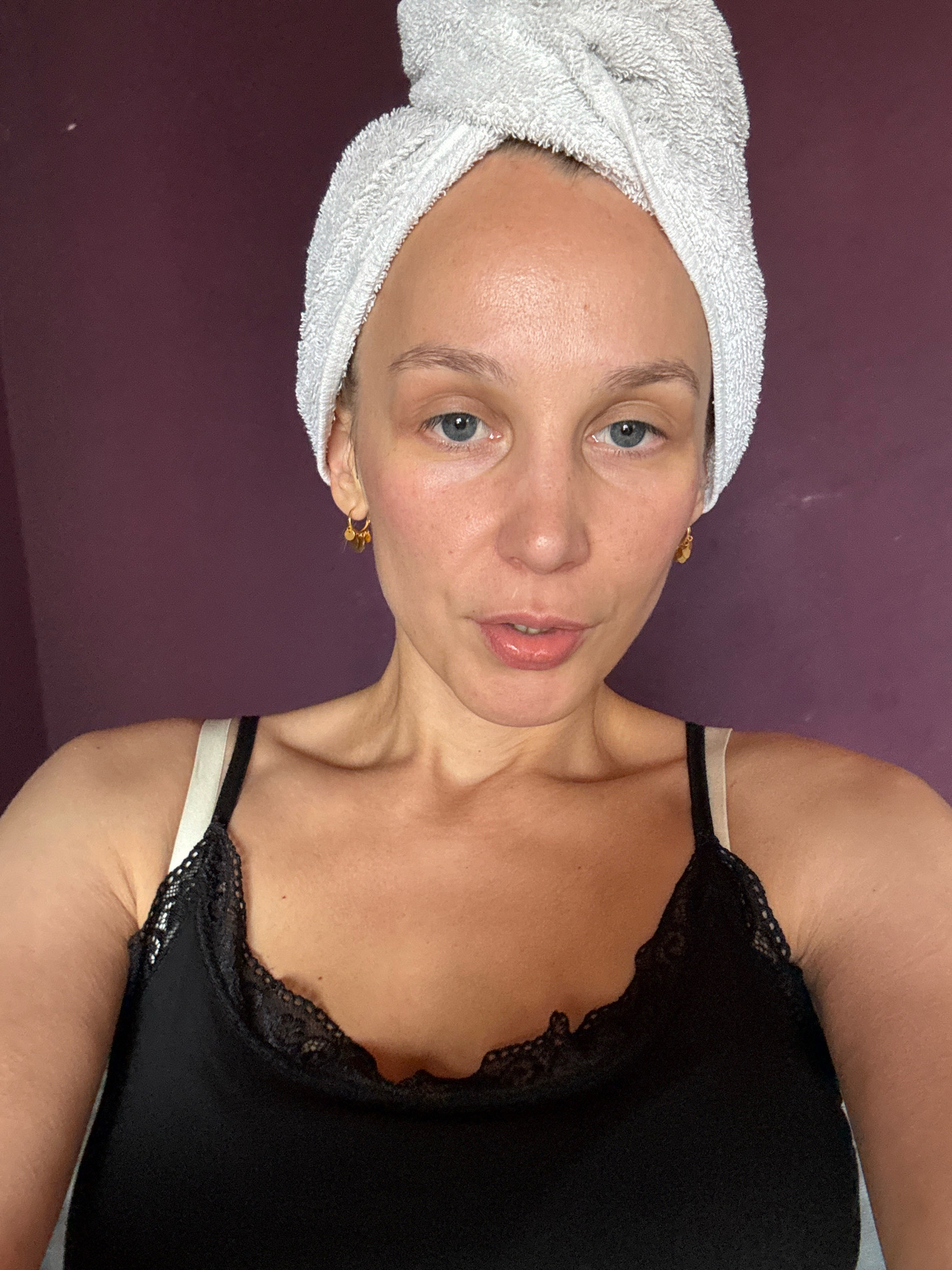 A person is taking a selfie indoors in front of a purple wall. The person has a white towel wrapped around their head and is wearing a black camisole with lace trim and white straps. They are looking directly at the camera with blue eyes and have light skin with some blush. There is a small gold earring in their visible ear. The lighting appears to be natural and coming from the front. The background is a solid purple wall. The scene does not provide any information indicating it is located in Überherrn, Germany.