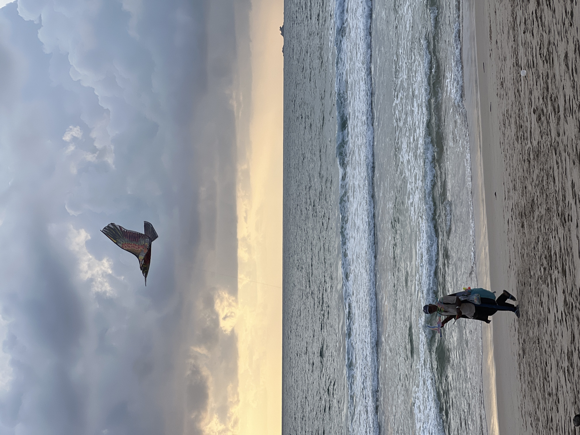 A man walks along a sandy beach at the edge of the sea. He is wearing a hat, a shirt, and dark pants, and carries a kite shaped like a bird in his left hand, along with a bag and other items. The kite is airborne in the sky, which is filled with dramatic, cloudy formations and a pale yellow band of light near the horizon. The ocean is calm with gentle waves. In the distance, a small island or landmass is visible on the horizon. The scene appears to be located in Choeng Thale, Thailand.