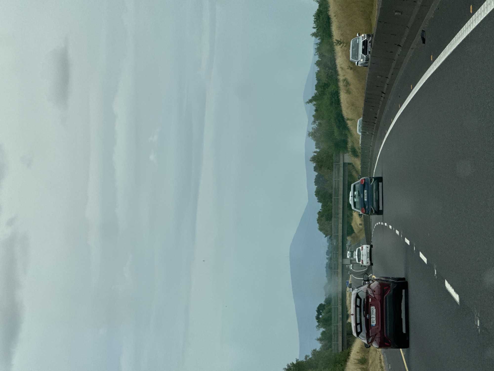The image captures a view from a vehicle traveling on a highway in Pill Bealin, Ireland. The road is asphalt with white lane markings. Several cars are visible, including a red hatchback in the foreground on the left, a white hatchback further ahead, and a dark blue SUV on the right. The landscape features rolling hills with lush green trees and dry grass. A bridge crosses the highway in the distance, and a mountain range is visible on the horizon under a cloudy sky. The overall atmosphere is overcast.