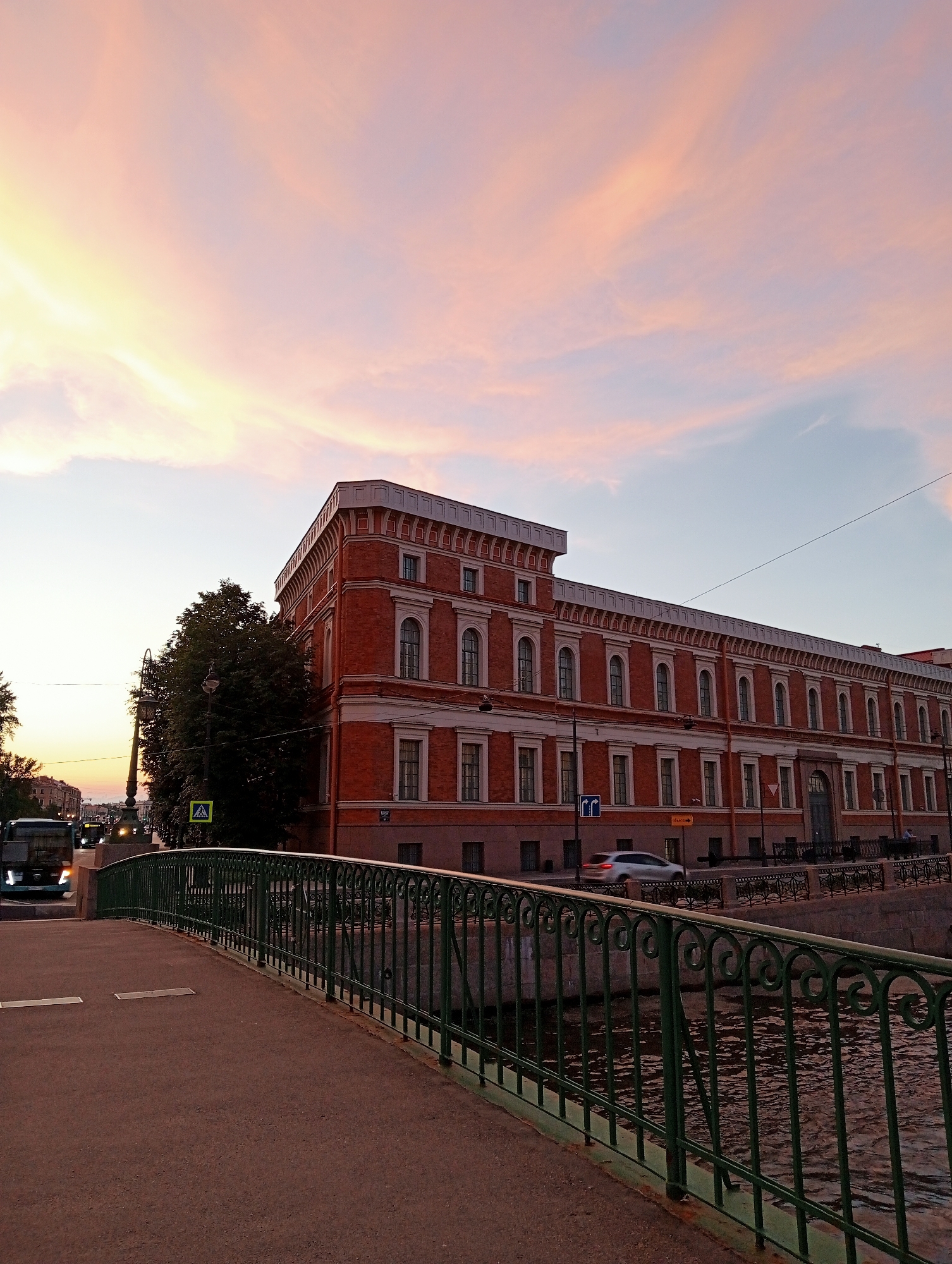 This is a photograph of a canal in Saint Petersburg, Russia, during a sunset. The sky is filled with soft pink and orange clouds, illuminated by the setting sun. In the foreground, a paved walkway leads over a bridge with a dark green metal railing. The railing has an ornate, decorative pattern of swirls and vertical bars. The canal water below reflects the muted colors of the sky. Across the canal, a long, red brick building with white trim and numerous arched windows stands prominently. A few trees with dark green foliage are visible to the left of the building. On the street behind the building, a silver car is in motion, and a bus is approaching from the left. Pedestrian crossing signs and streetlights are also present. The overall scene captures a tranquil urban landscape at dusk.