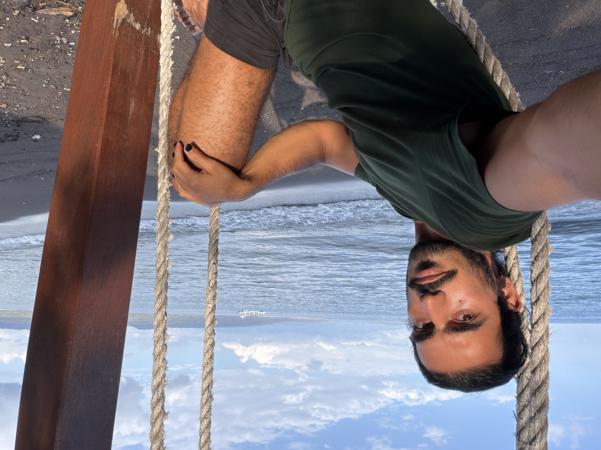 In Unknown, Indonesia, a man with dark hair and a beard sits on a swing by the ocean. He wears a green t-shirt and dark shorts. The swing is made of thick rope and has a wooden support. The background shows a calm sea with a small boat in the distance, and a cloudy sky. The beach is dark sand. The man is looking towards the camera, and his left arm is resting on his knee. The image appears to be a selfie, taken at eye level.